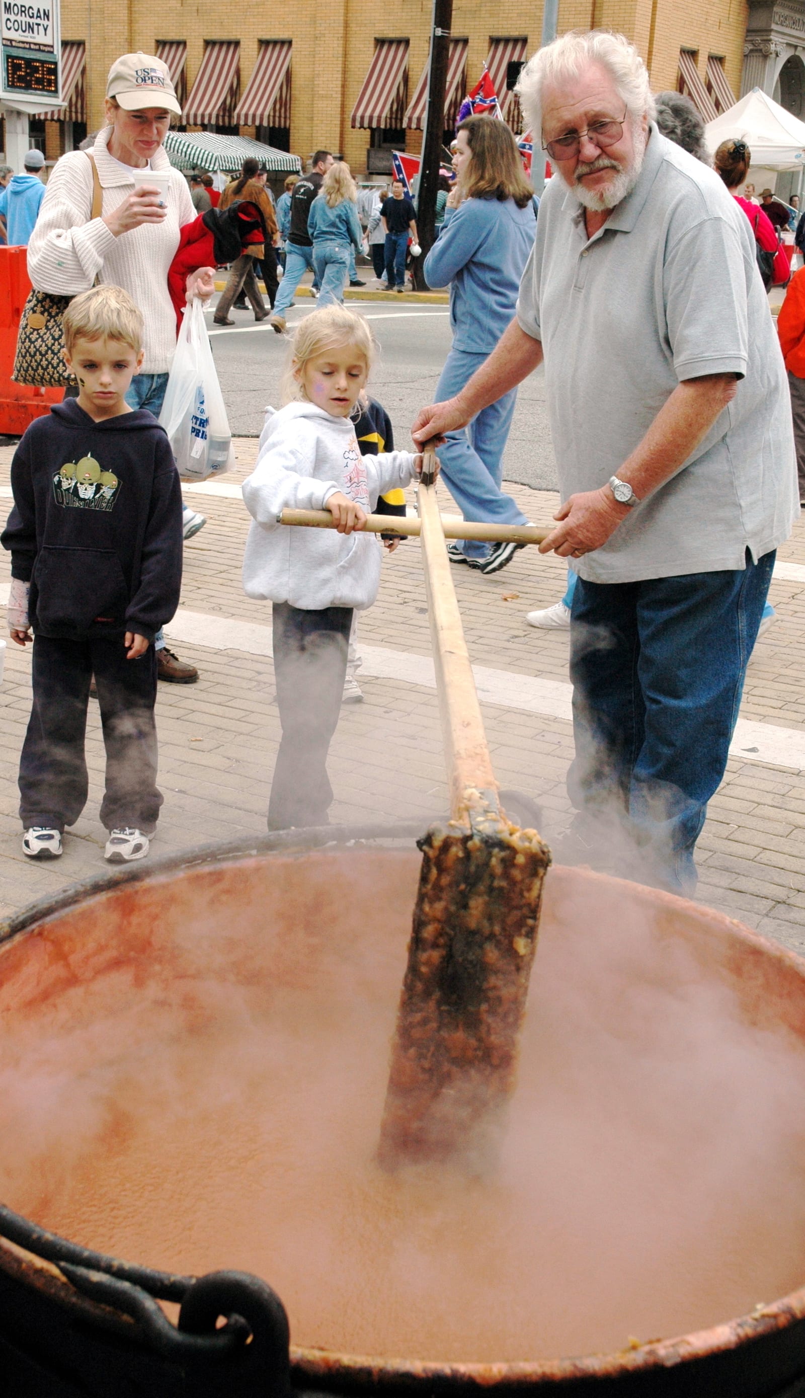 A child and an older man stirring steaming apple butter in a large copper kettle at a street festival.