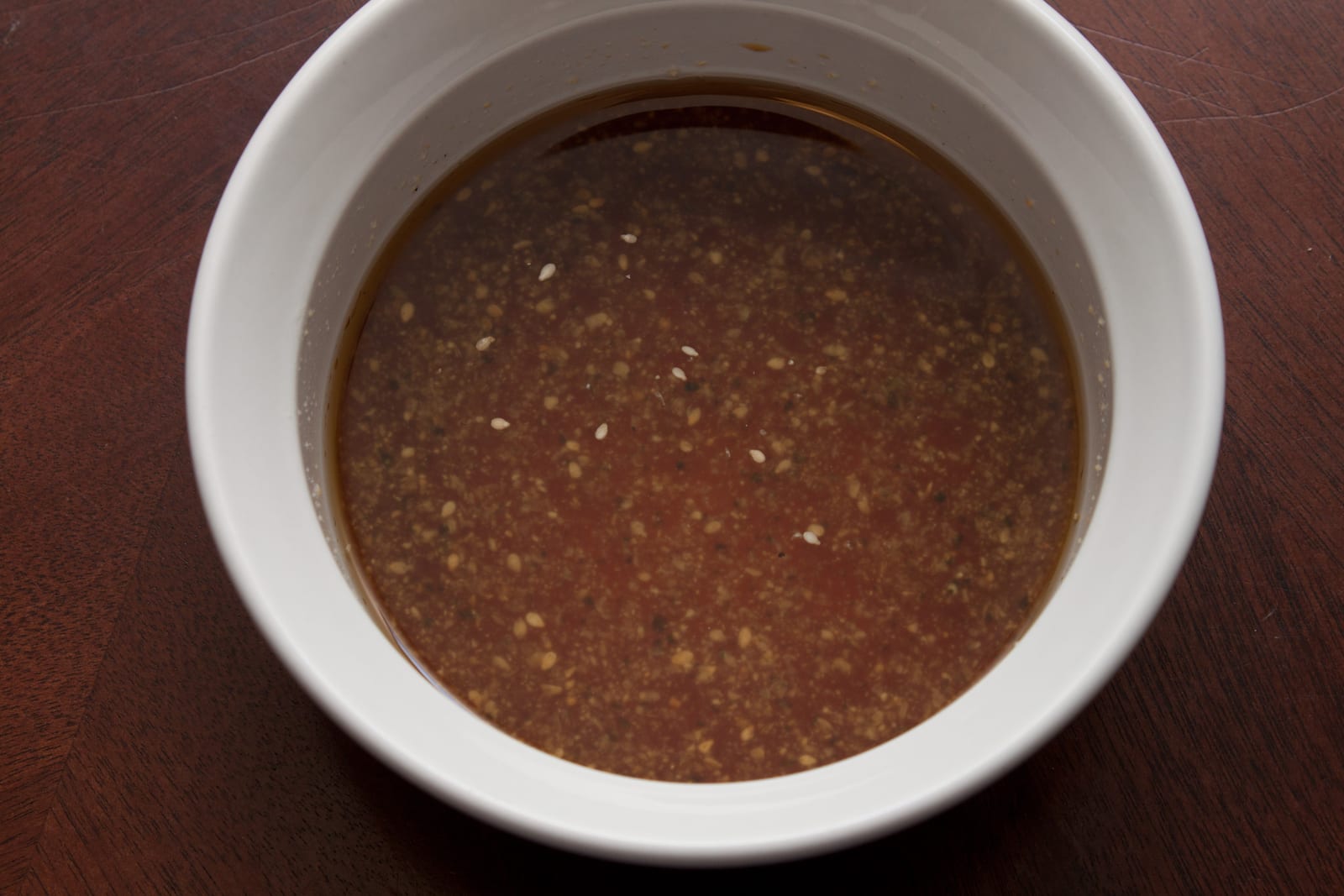 A thin brown sesame dressing flecked with seasoning in a white bowl.