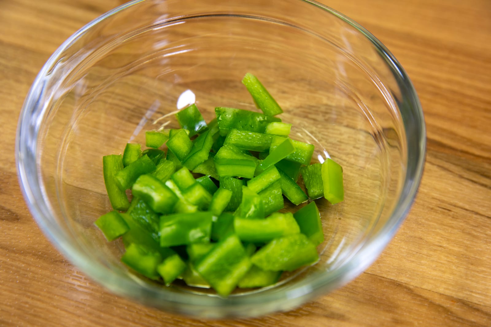Chopped green bell pepper in a clear glass bowl on a wooden surface.