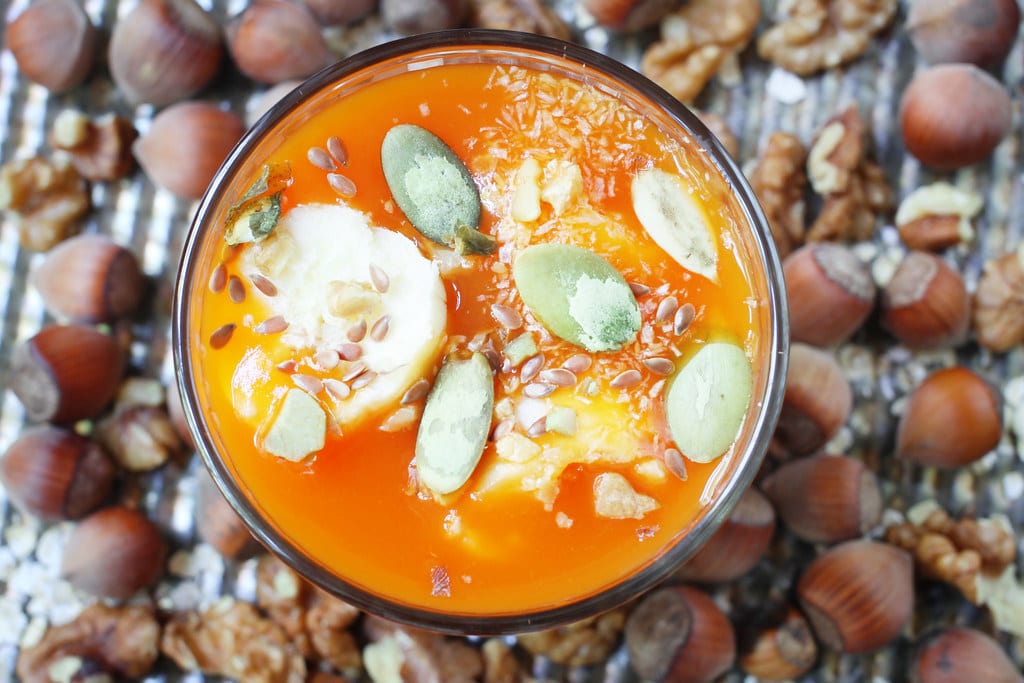 A vivid orange pumpkin smoothie in a bowl, topped with pumpkin seeds and surrounded by hazelnuts.