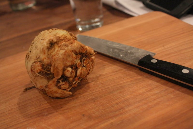 A small whole celeriac root beside a chef’s knife on a wooden board.