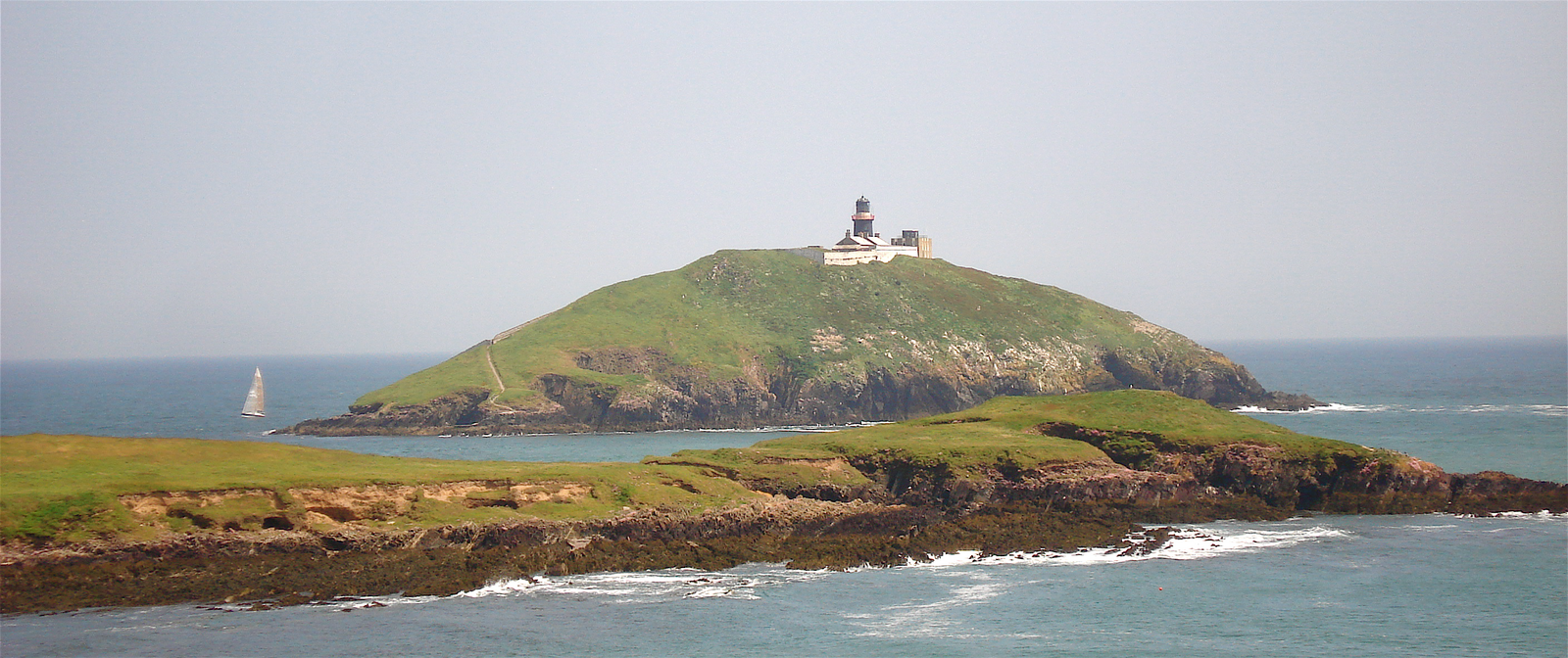 Ballycotton Lighthouse on a green island beyond a rocky County Cork shoreline.
