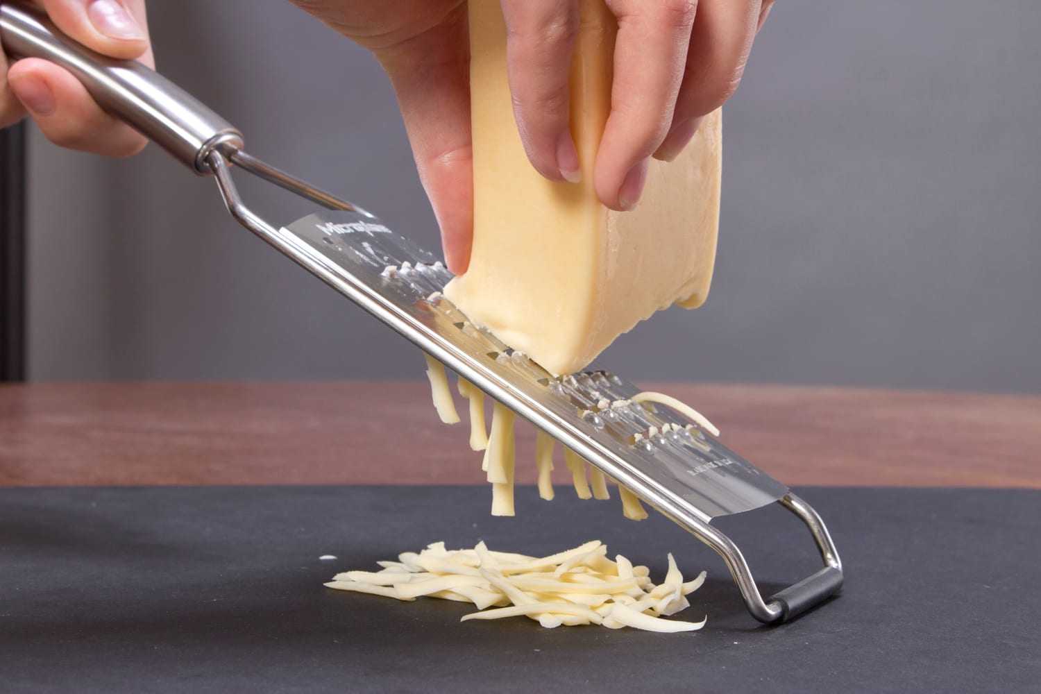 Hands drawing a pale block of cheese across a flat grater over a dark work surface.