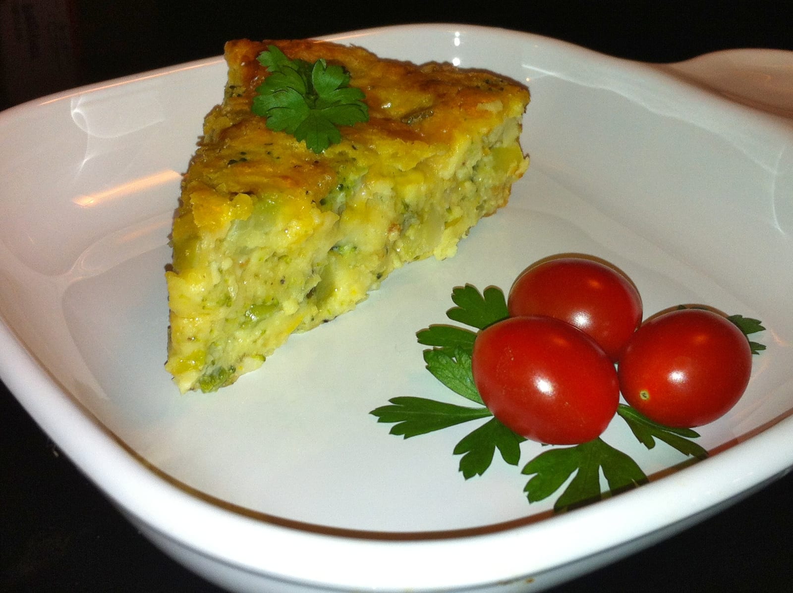 A thick slice of broccoli quiche beside cherry tomatoes and parsley.