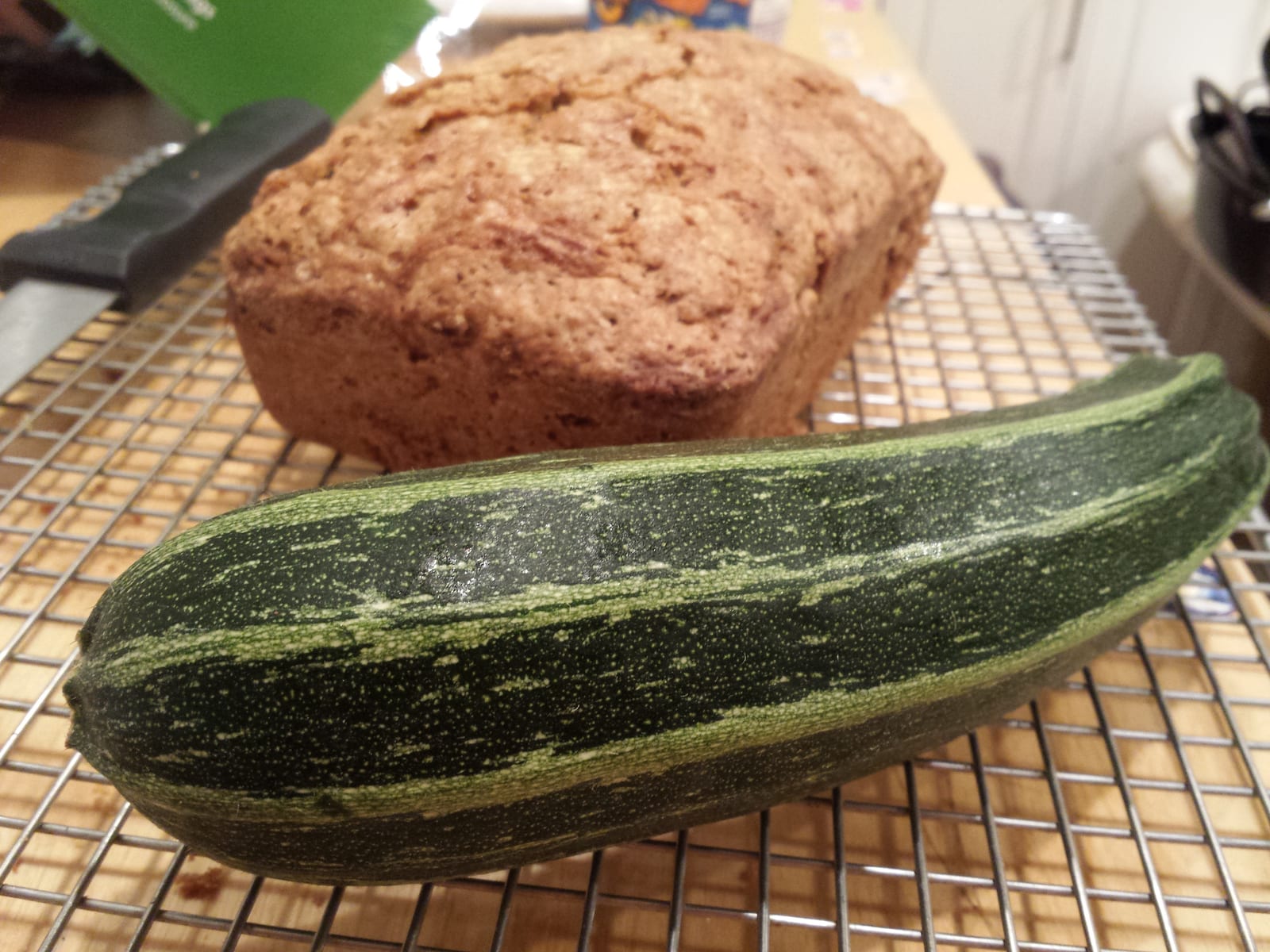 A large striped zucchini in front of a cooling loaf of zucchini bread.