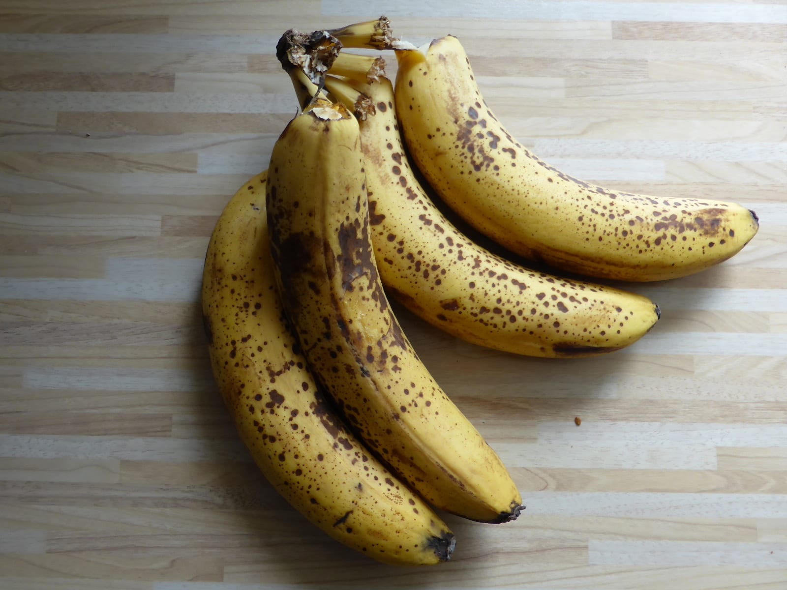 Four ripe yellow bananas covered with brown freckles on a wooden worktop.