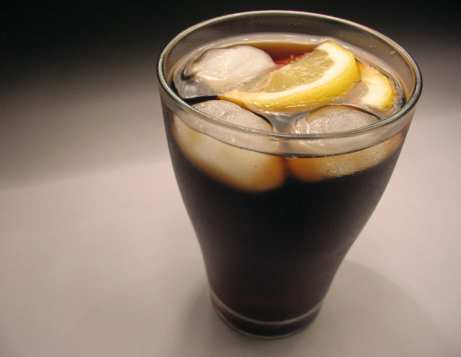 A cold glass of dark cola with ice cubes and lemon slices against a dark background.