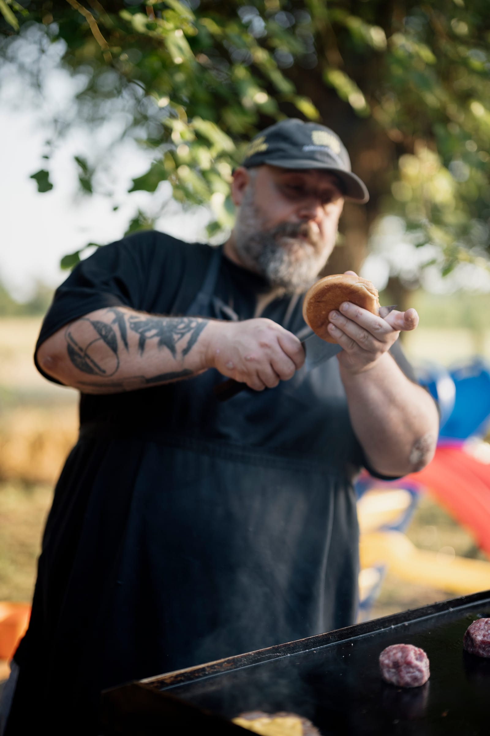 A cook assembling a burger beside patties on a smoking outdoor flat-top grill.