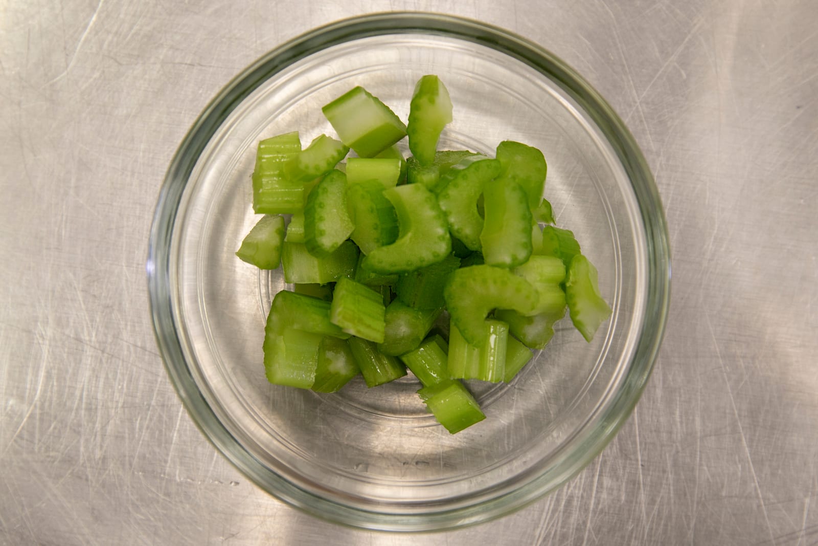 Chopped green celery in a small glass bowl on a stainless-steel surface.