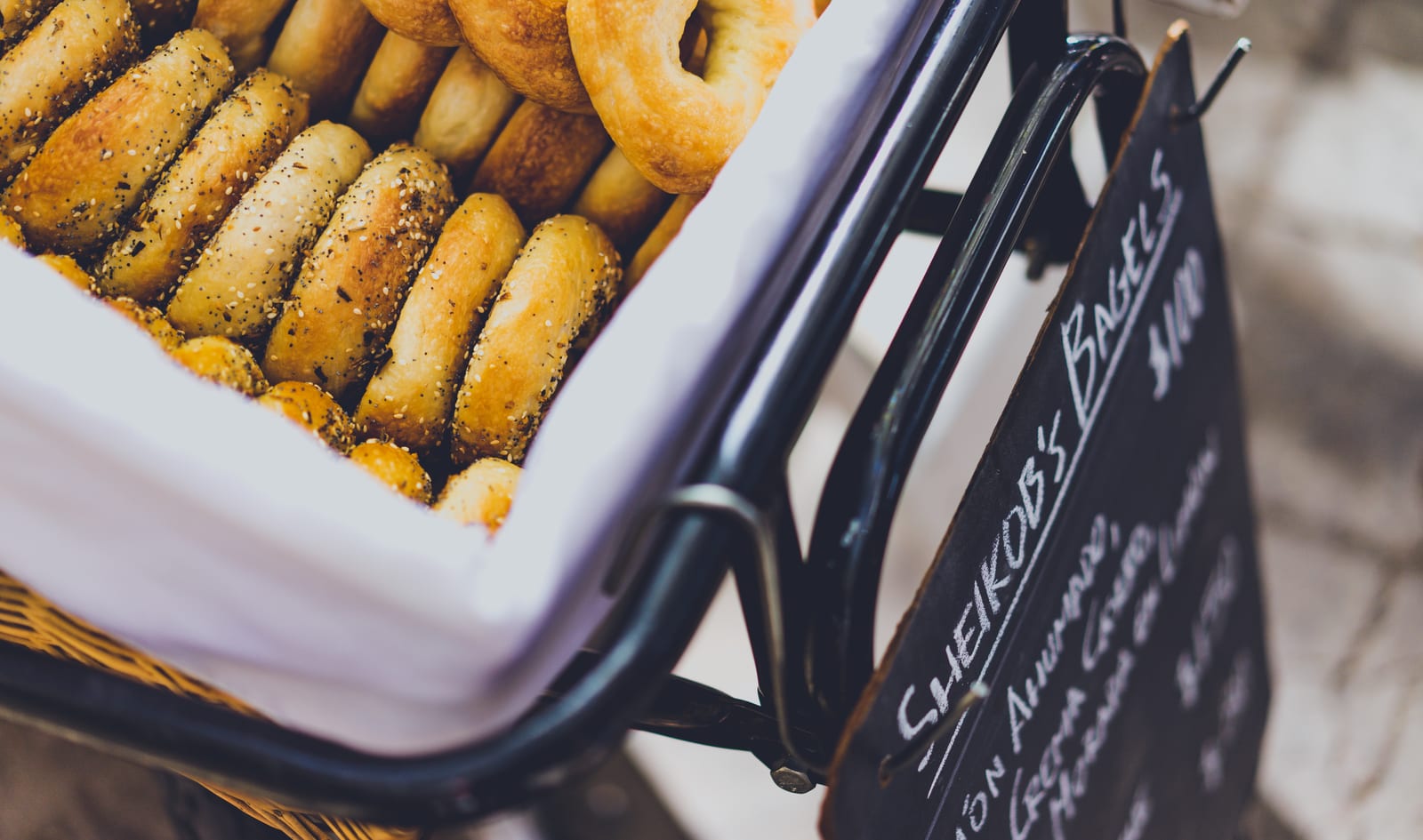Fresh plain and seeded bagels piled in a market basket beside a chalkboard sign.