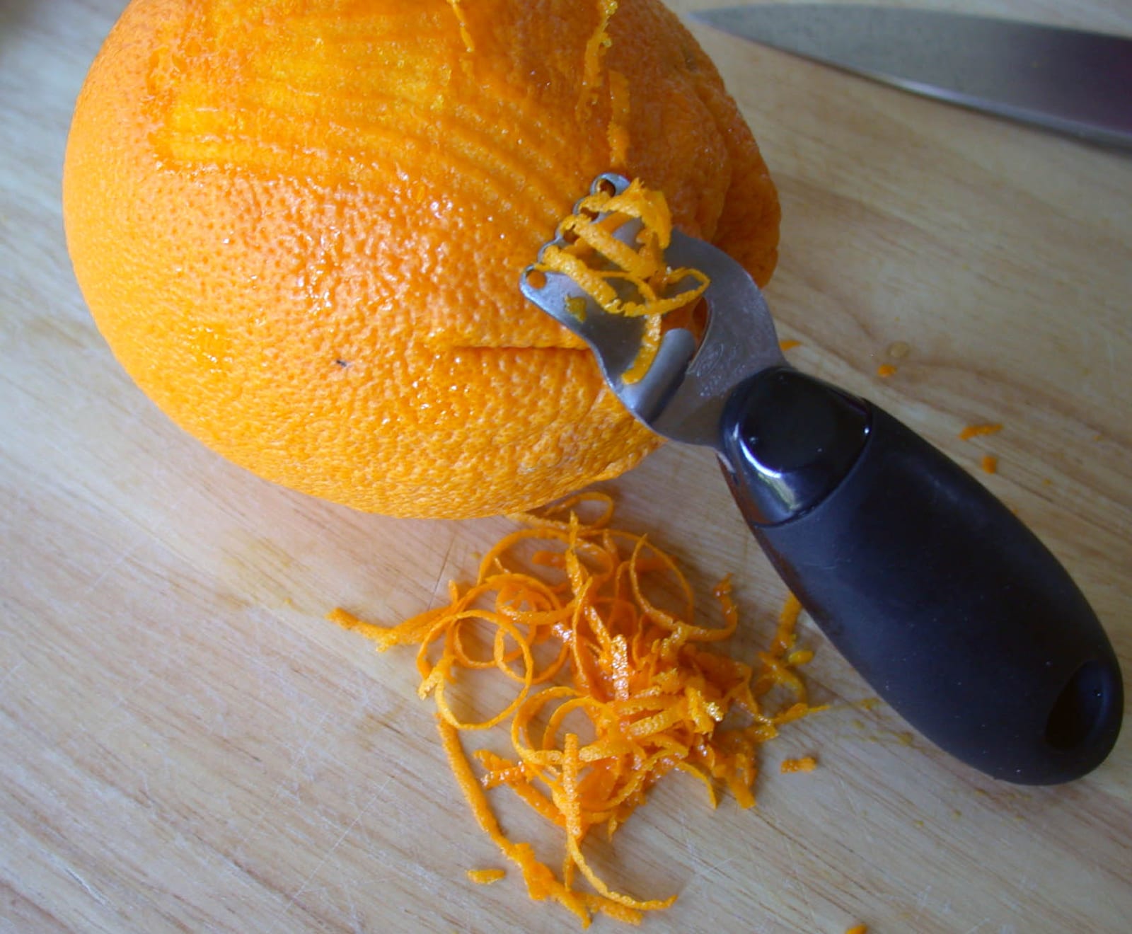 A hand zester drawing bright strips of peel from a whole orange.