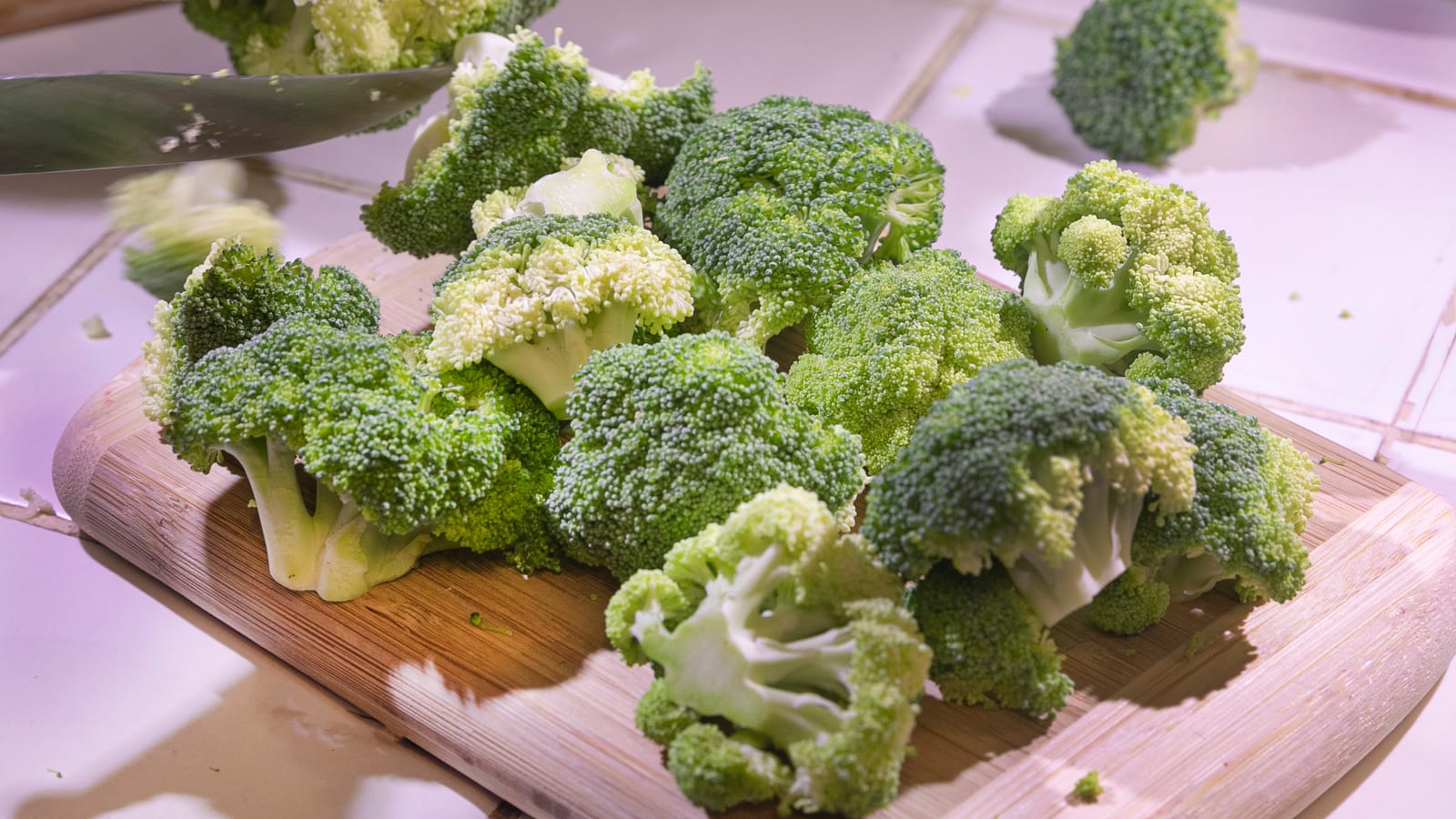 Fresh broccoli florets being cut on a wooden board with a chef’s knife.