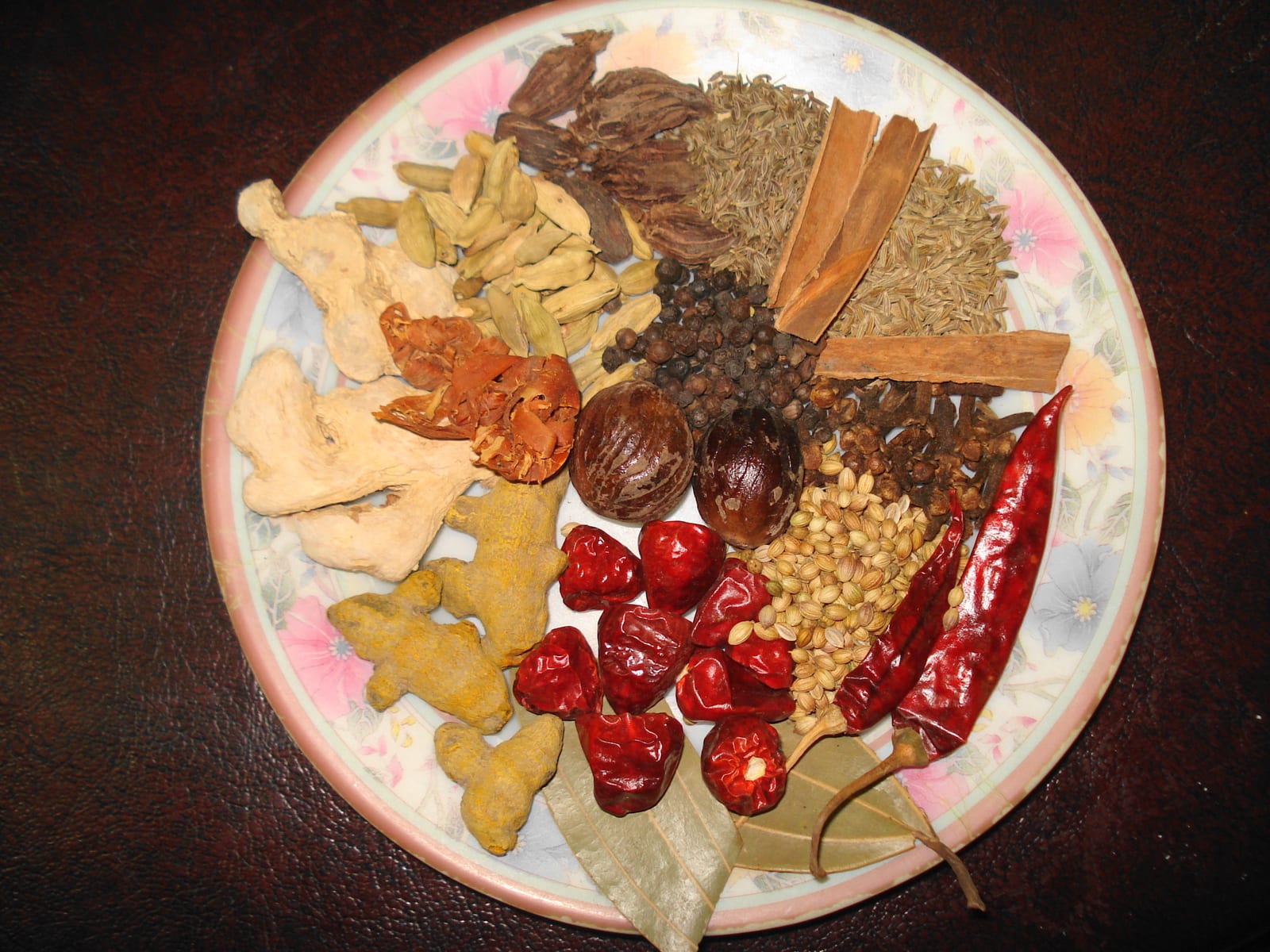 Whole dried spices including ginger, turmeric, cardamom, cumin, coriander, cinnamon, pepper, chiles, cloves, and bay leaves arranged on a plate.