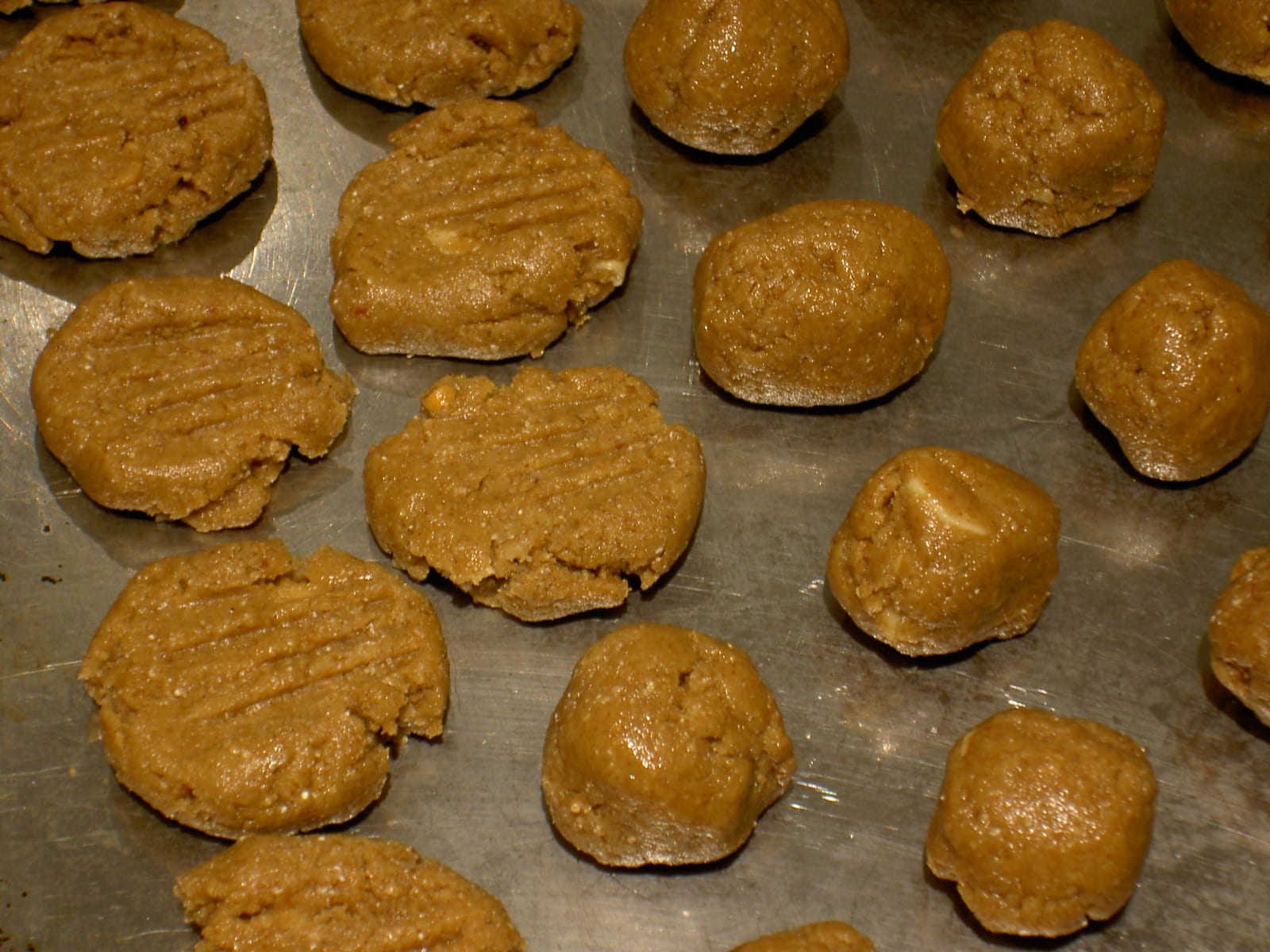 Portioned peanut butter cookie dough on a metal baking sheet, with some pieces pressed flat.
