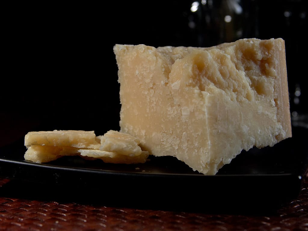 A wedge of aged Parmigiano-Reggiano with several thin shards on a dark plate.