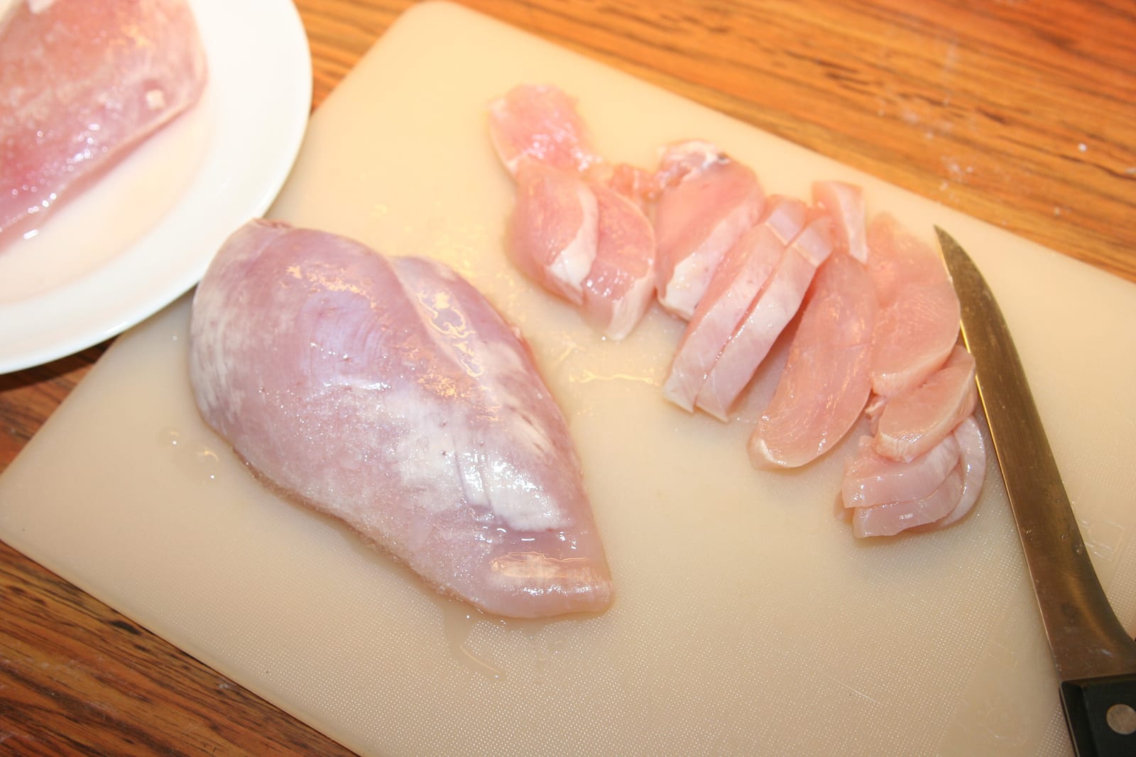 Raw boneless chicken breasts being sliced into strips on a white cutting board.
