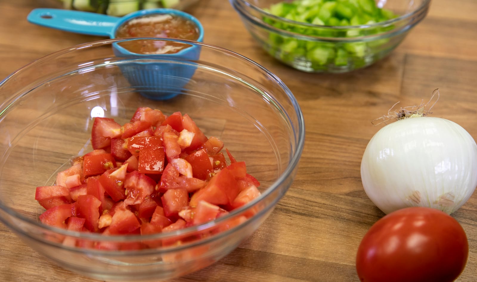 Diced tomatoes in a glass bowl beside whole tomatoes, onion, celery, and a small cup of sauce.