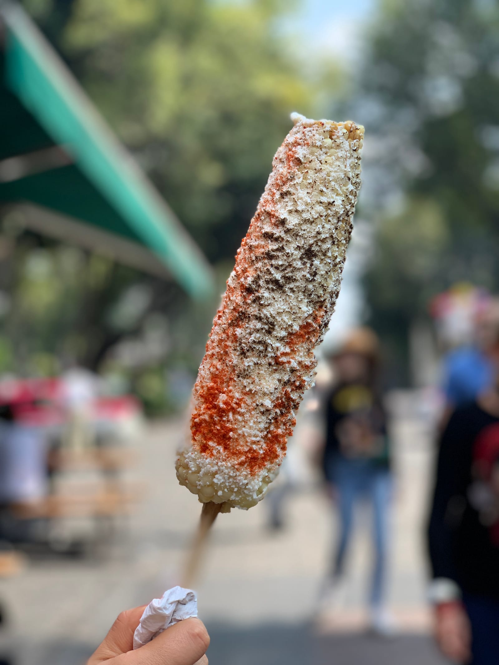 A hand holding a cheese- and chile-covered elote on a wooden stick at a street stand in Mexico.