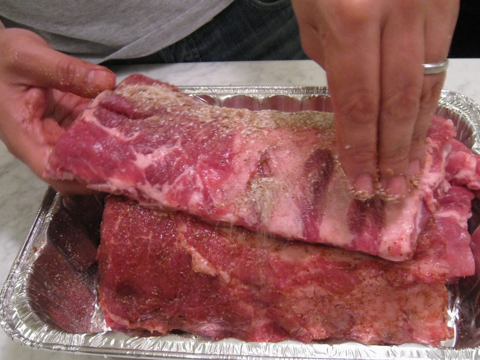 Hands applying seasoning to raw pork ribs in a foil pan.