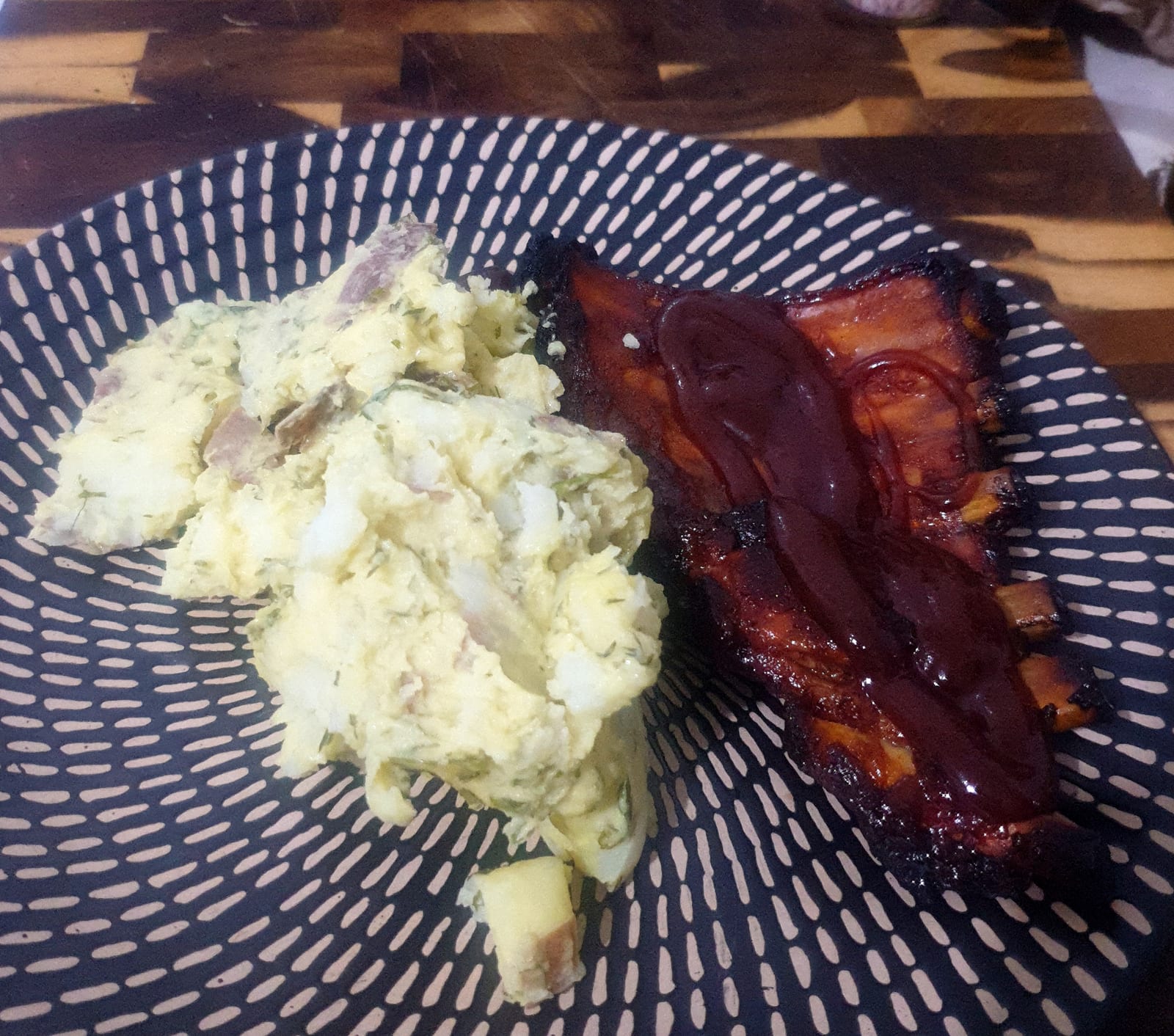 Barbecue pork ribs with extra sauce served beside potato salad in a patterned bowl.