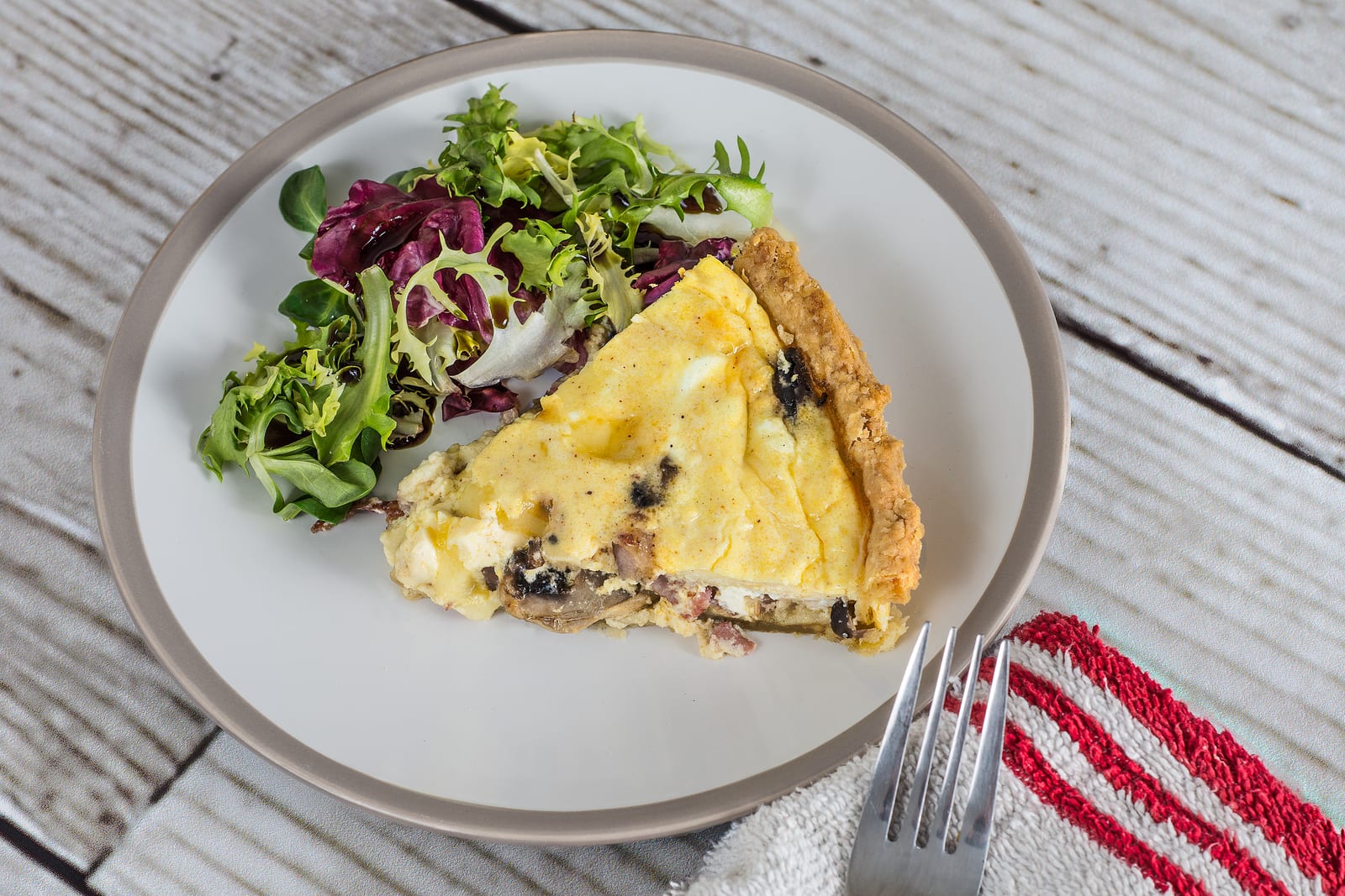 A slice of mushroom quiche beside dressed salad leaves on a white plate.