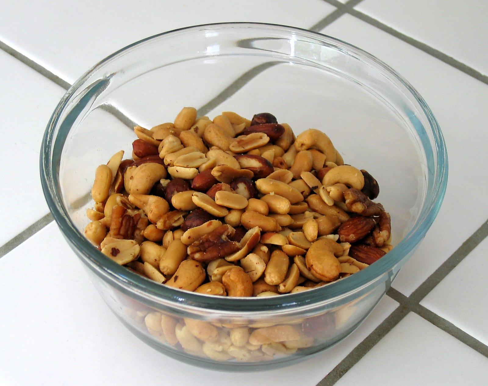 Roasted peanuts, cashews, almonds, pecans, and other mixed nuts in a clear glass bowl.