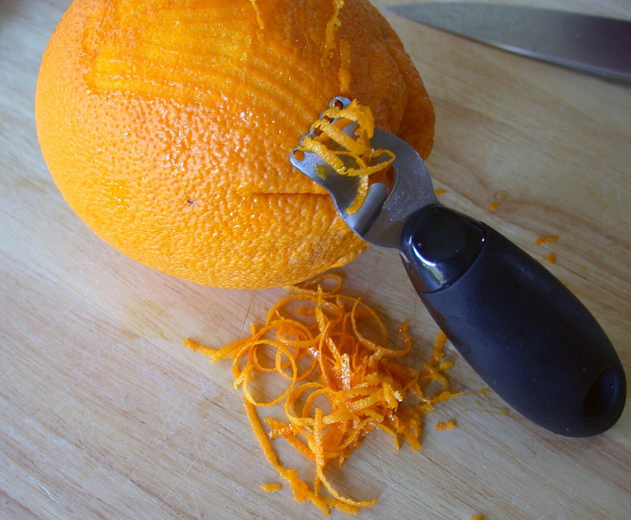 A hand zester cutting fine orange peel over a wooden board.