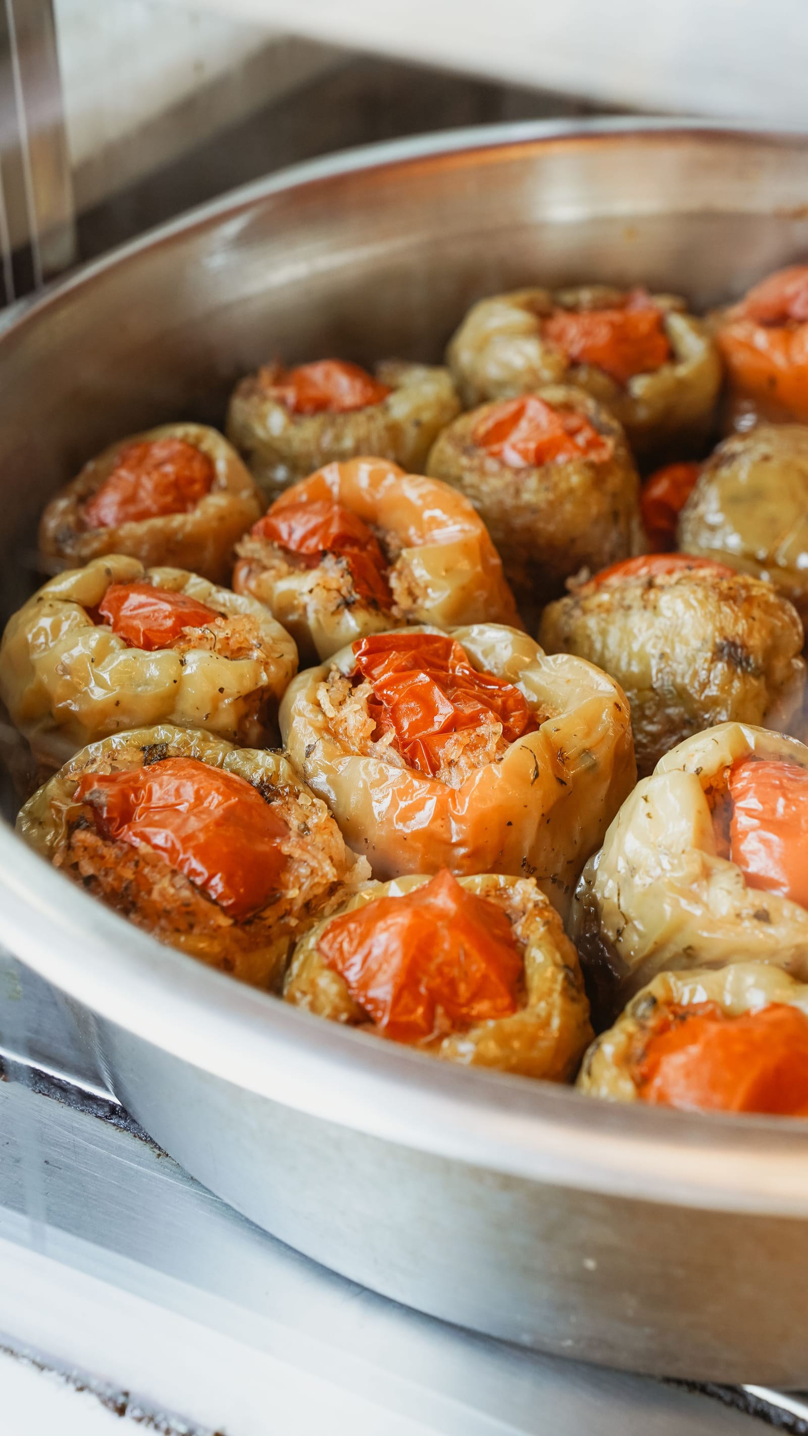A large metal pan crowded with soft whole peppers filled with rice and capped with tomatoes.