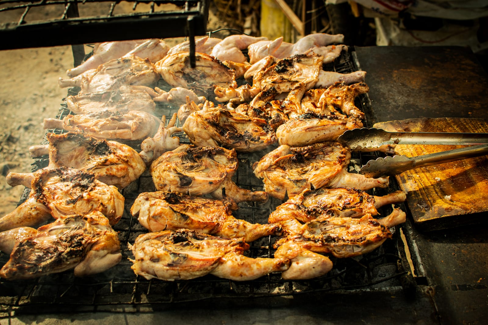 Many split chickens browning over a smoky open charcoal grill.