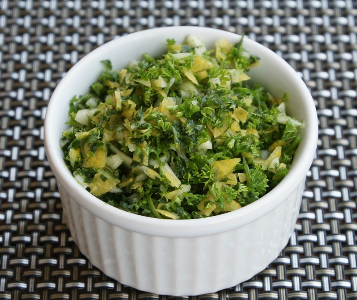 A small white ramekin filled with chopped parsley, garlic, and pieces of lemon peel.