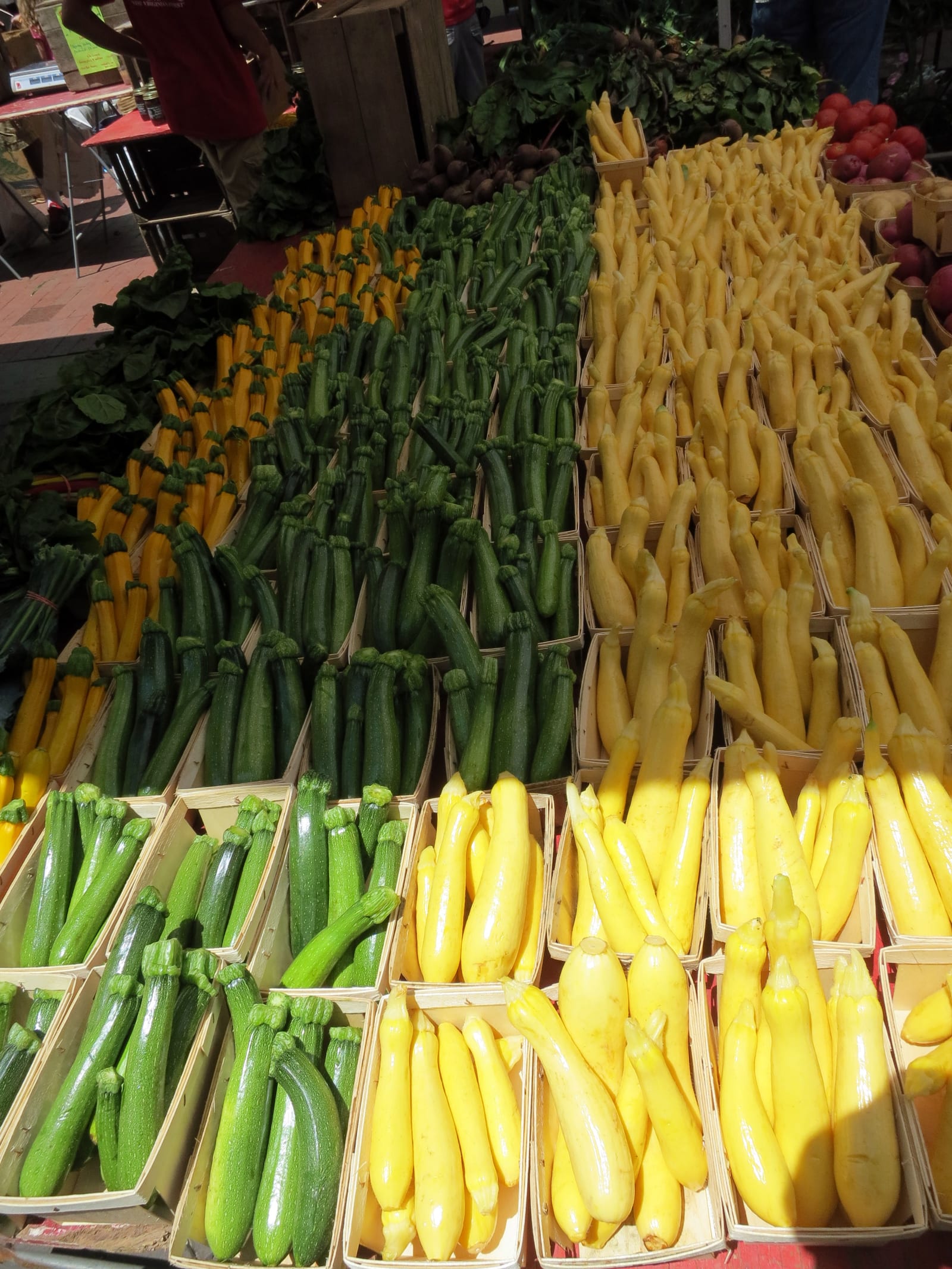 Green zucchini and yellow summer squash arranged in long rows of wooden baskets at a market.