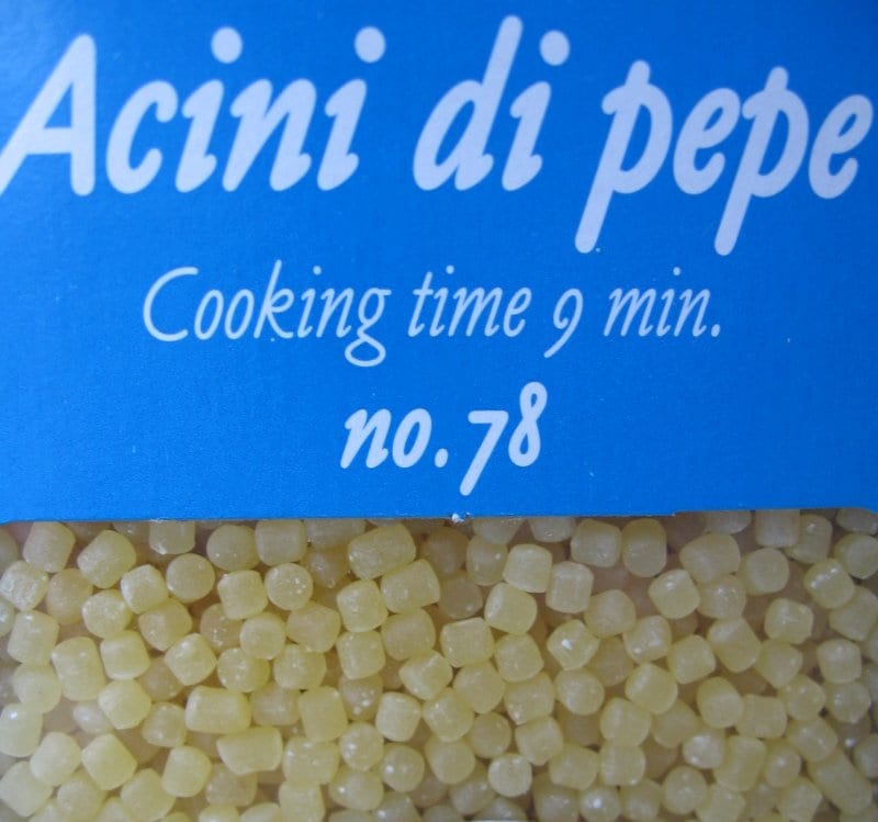 Tiny pale acini di pepe pasta visible through a blue pasta package window.