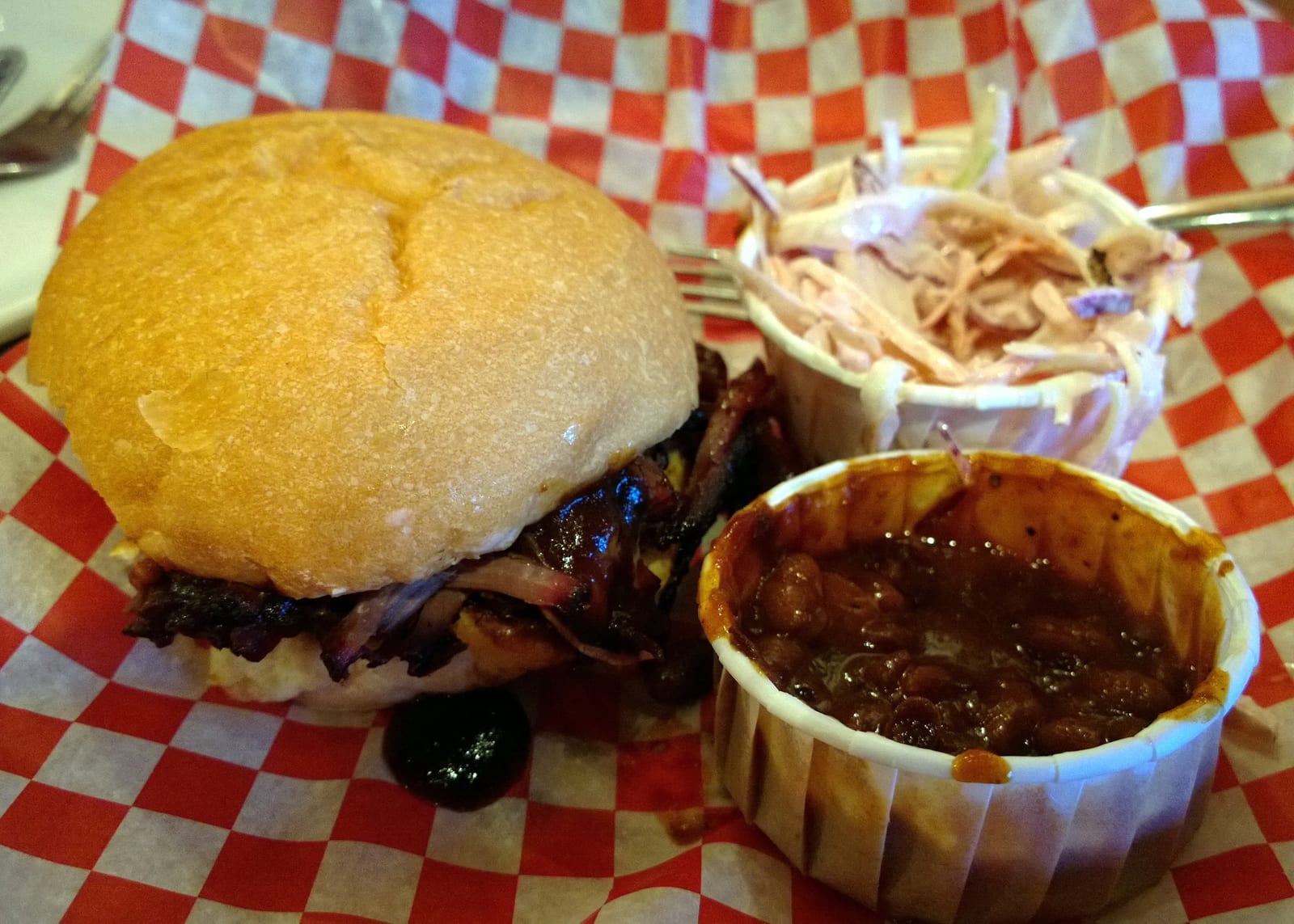 A brisket sandwich with a paper cup of barbecue beans and coleslaw on red checked paper.