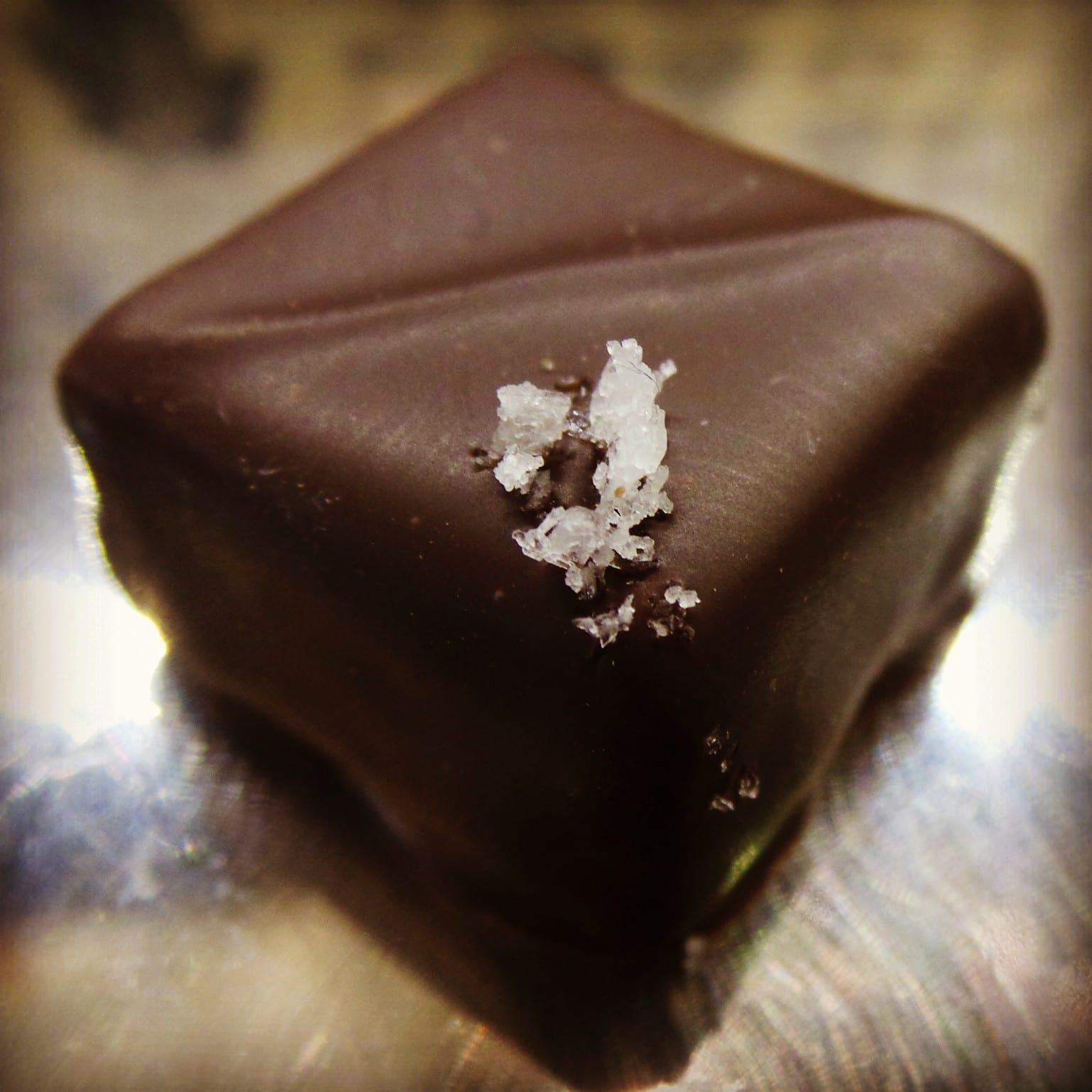 A glossy square of dark chocolate ganache topped with coarse salt crystals on a metal surface.