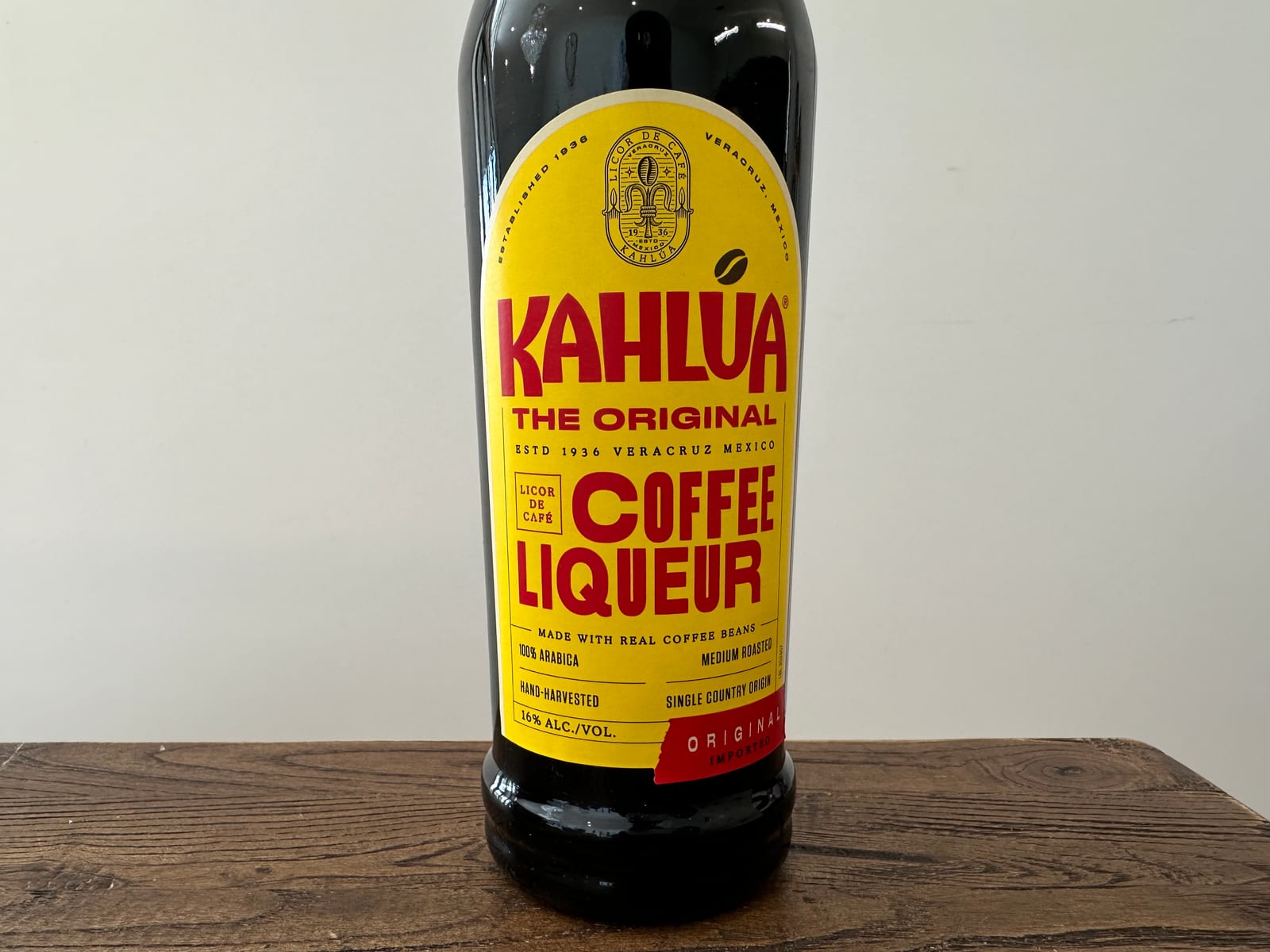A bottle of Kahlúa coffee liqueur standing on a wooden surface against a pale wall.