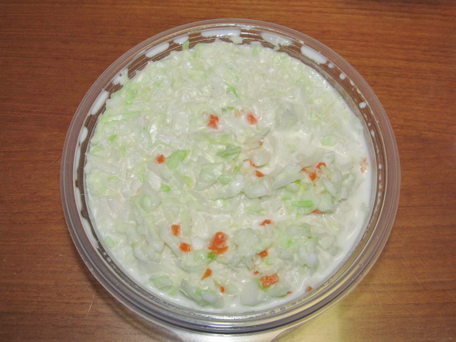 Finely chopped creamy coleslaw in a clear round restaurant container on a wood table.