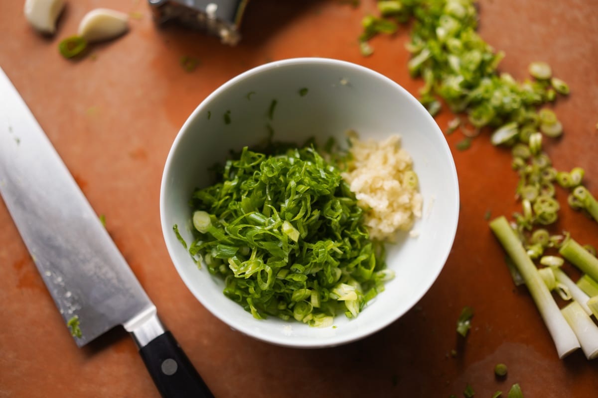 Finely sliced green onions and chopped garlic in a white bowl on a cutting board.