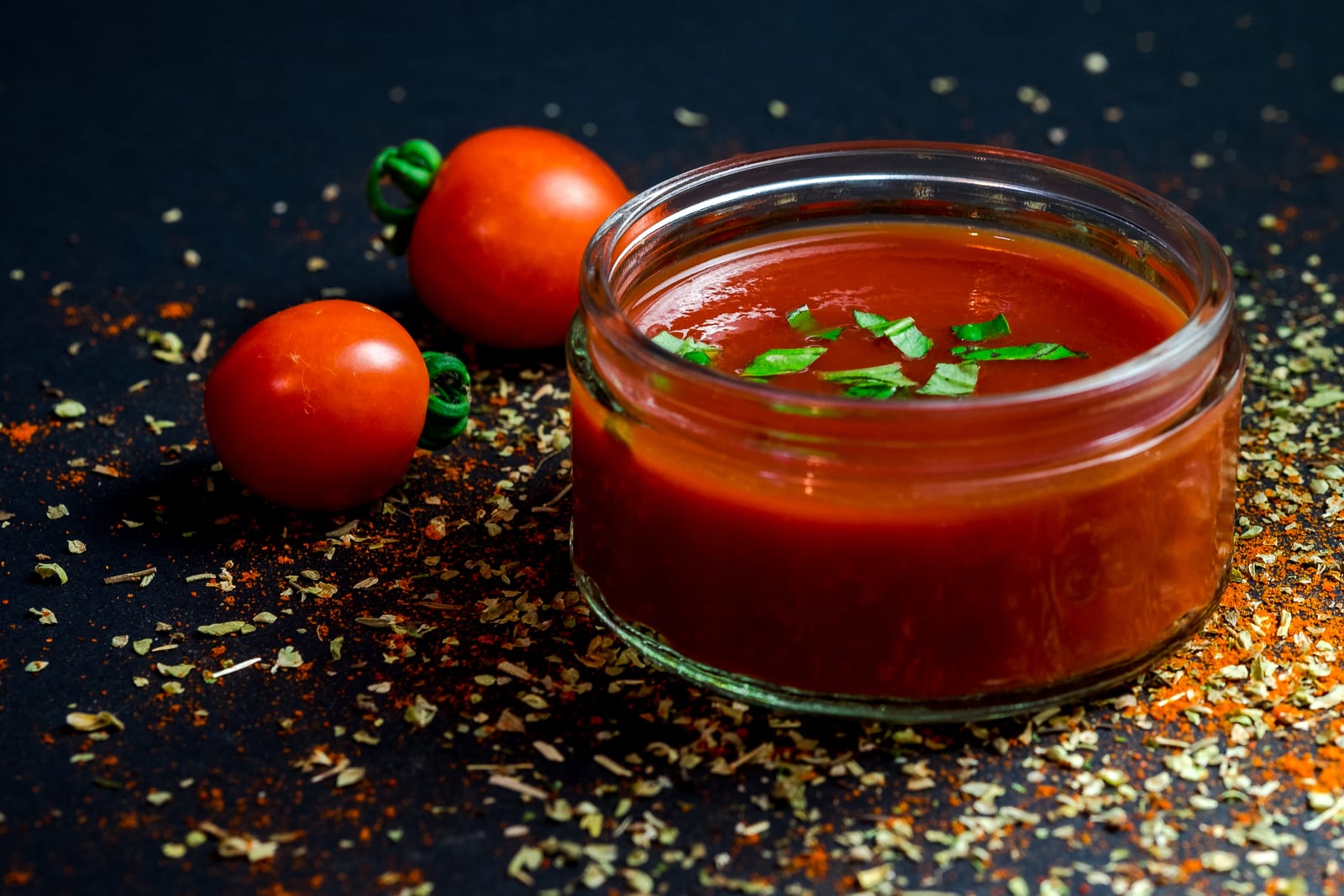 Bright red tomato sauce in a glass jar beside whole tomatoes and dried herbs.