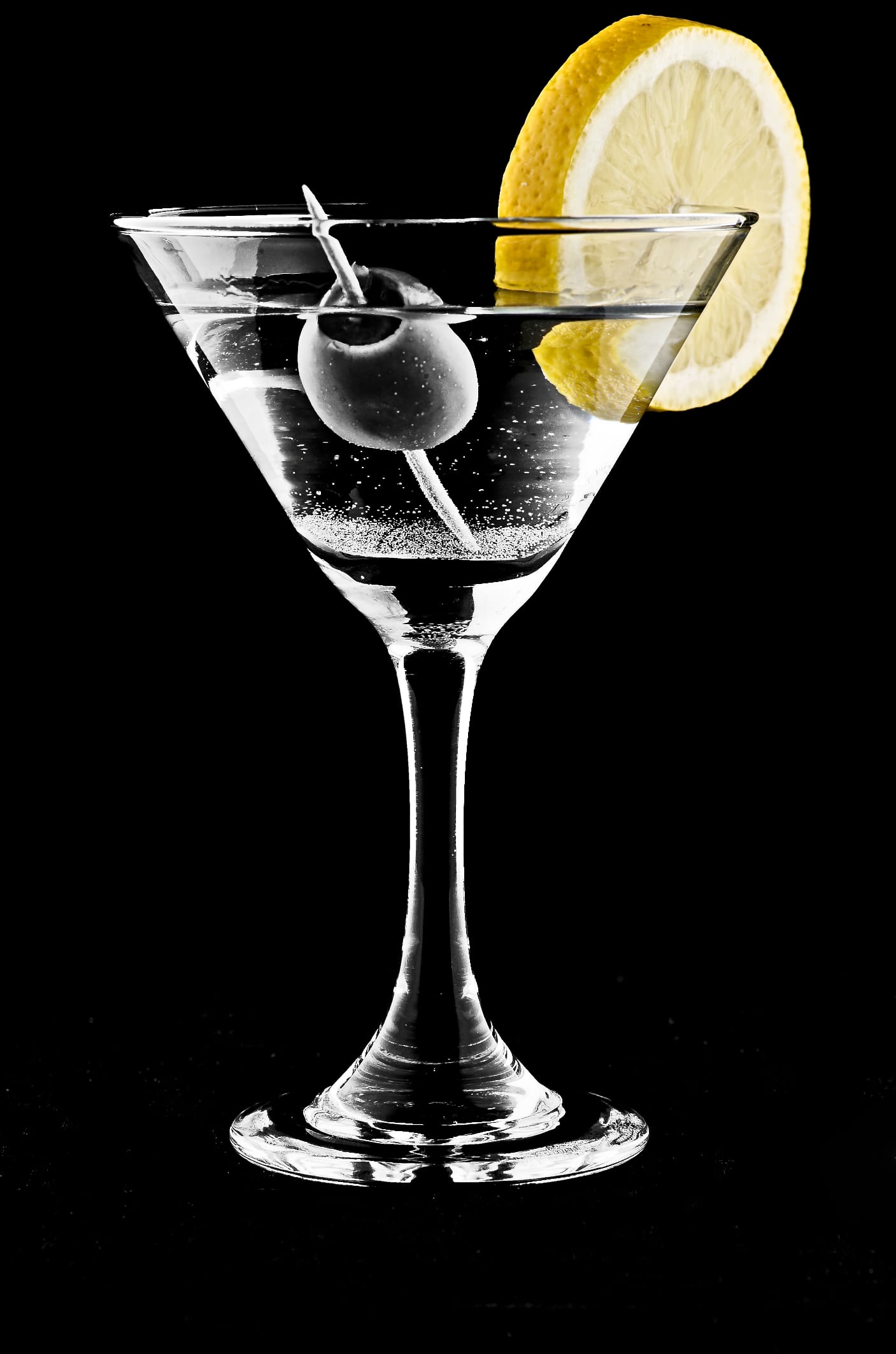 A clear vodka martini in a stemmed glass against a black background, garnished with lemon and an olive.