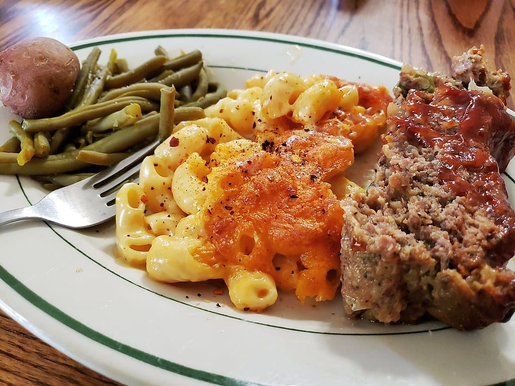 A homestyle dinner plate with meatloaf, macaroni and cheese, green beans, and a small potato.