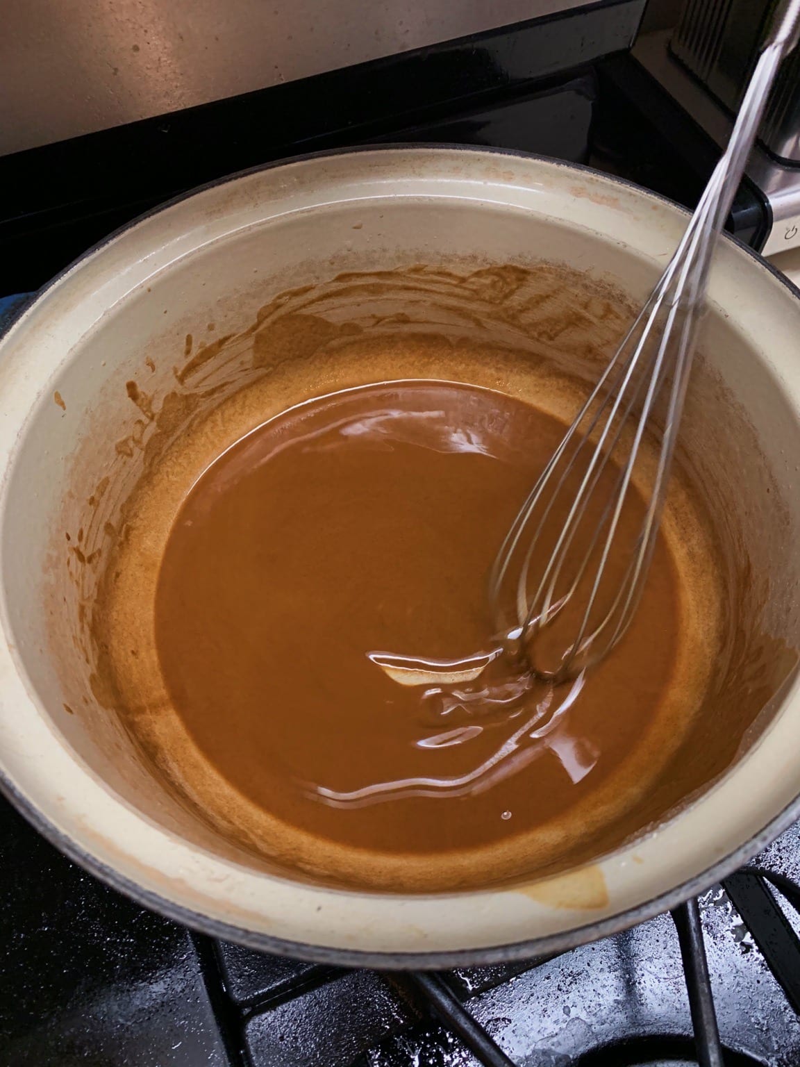A whisk resting in a glossy brown roux inside a pale enameled Dutch oven.