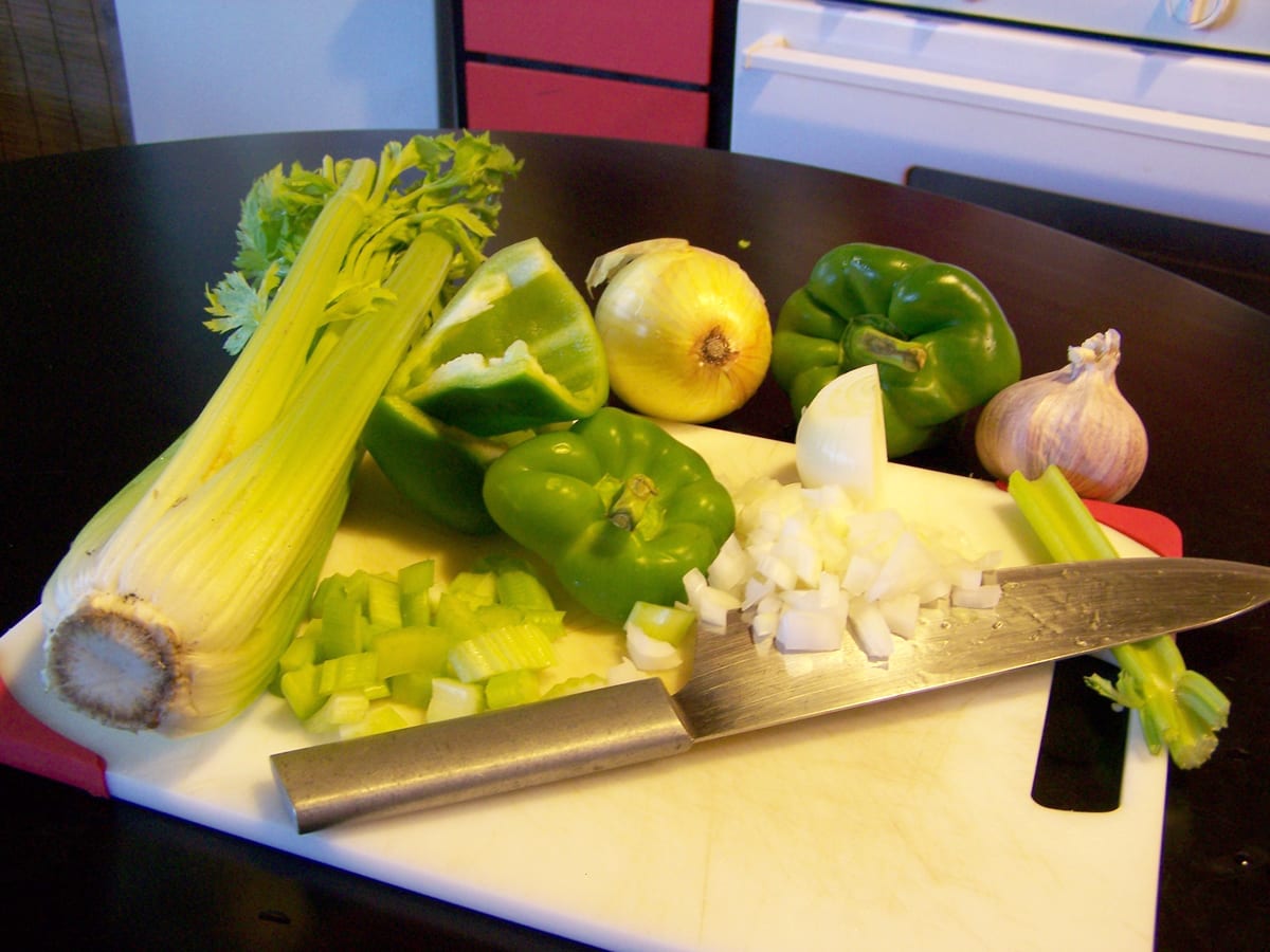 Celery, green bell peppers, yellow onions, and garlic arranged on a cutting board with a chef’s knife.