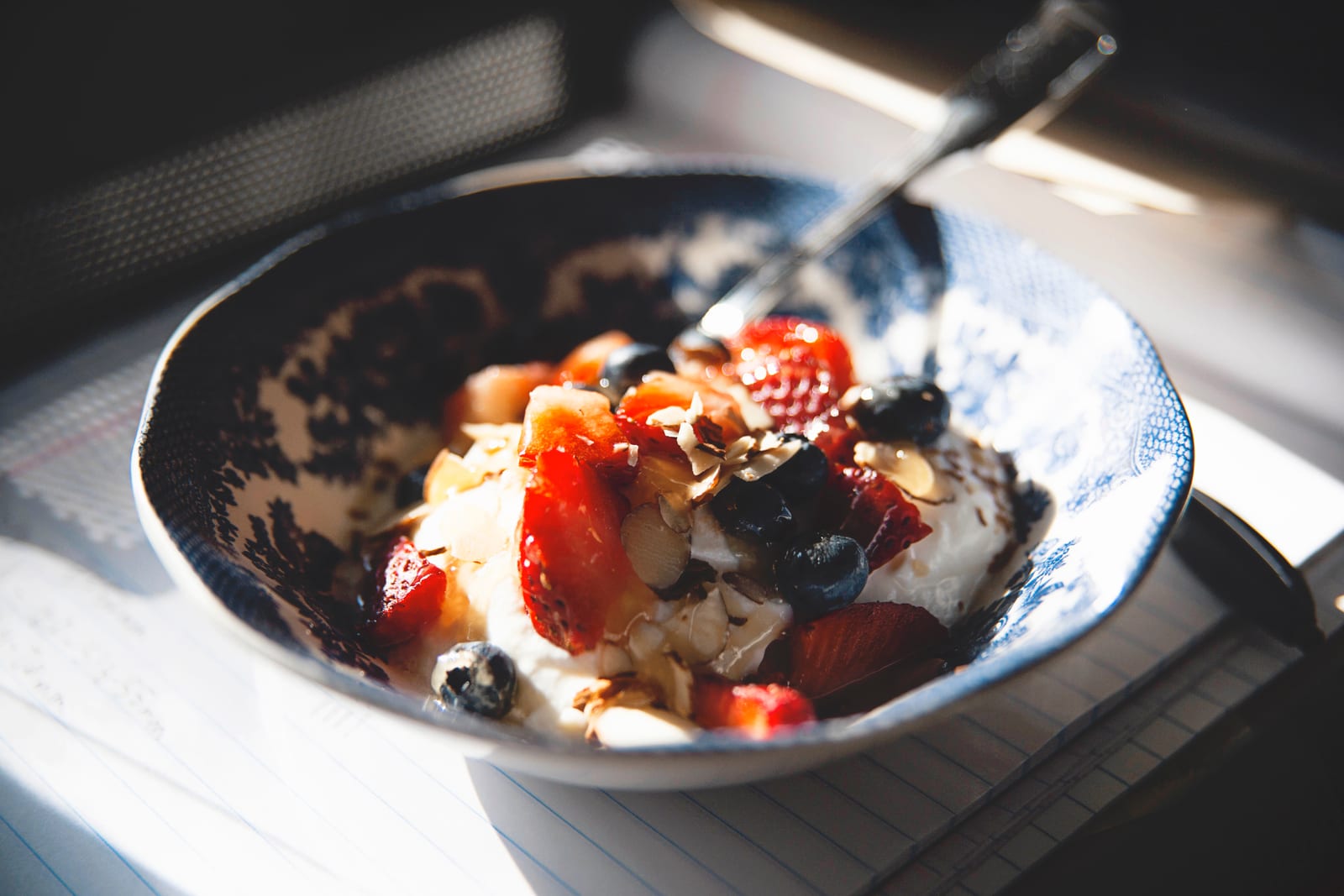 Thick yogurt topped with berries and sliced almonds in a blue-and-white bowl in window light.