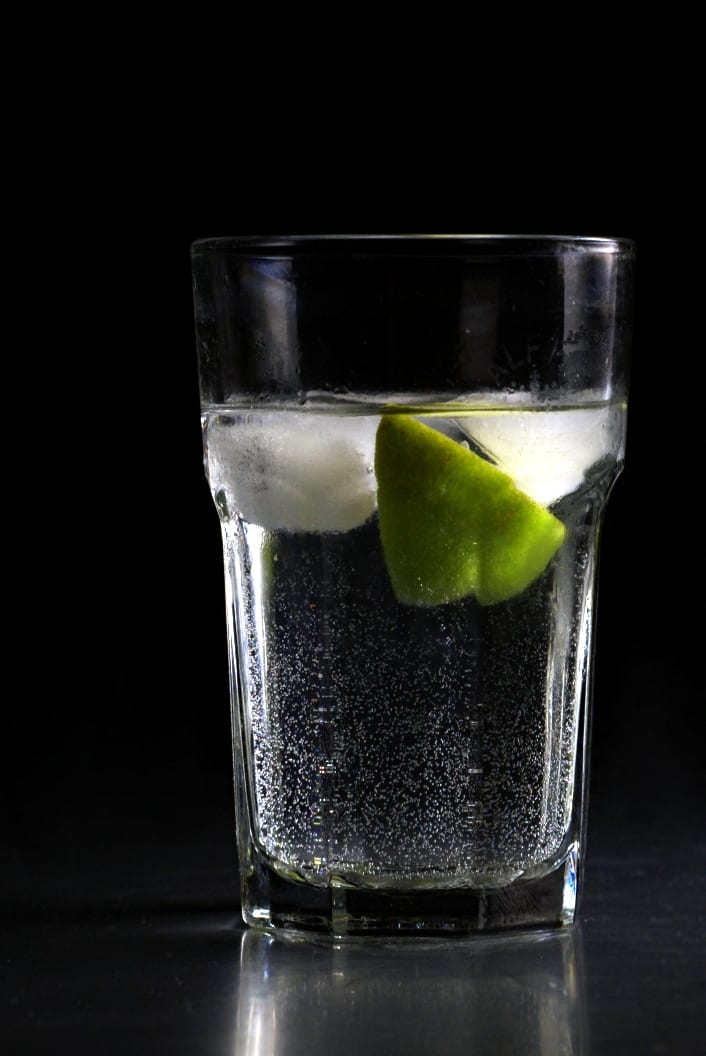 A tall highball glass filled with a clear sparkling drink, ice, and a lime wedge.