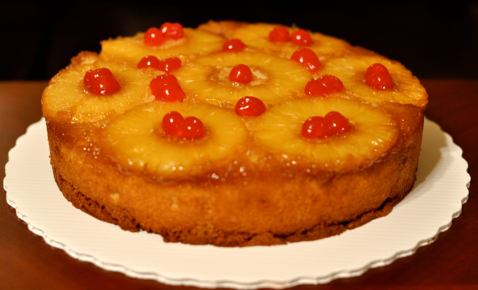 A round pineapple upside-down cake with browned pineapple rings and red cherries.