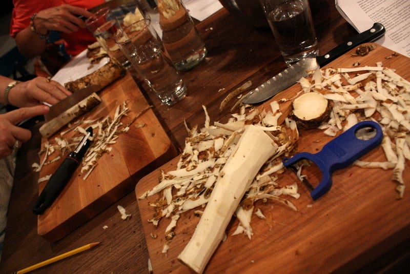 A fresh horseradish root being peeled on a wooden cutting board during a cooking class.