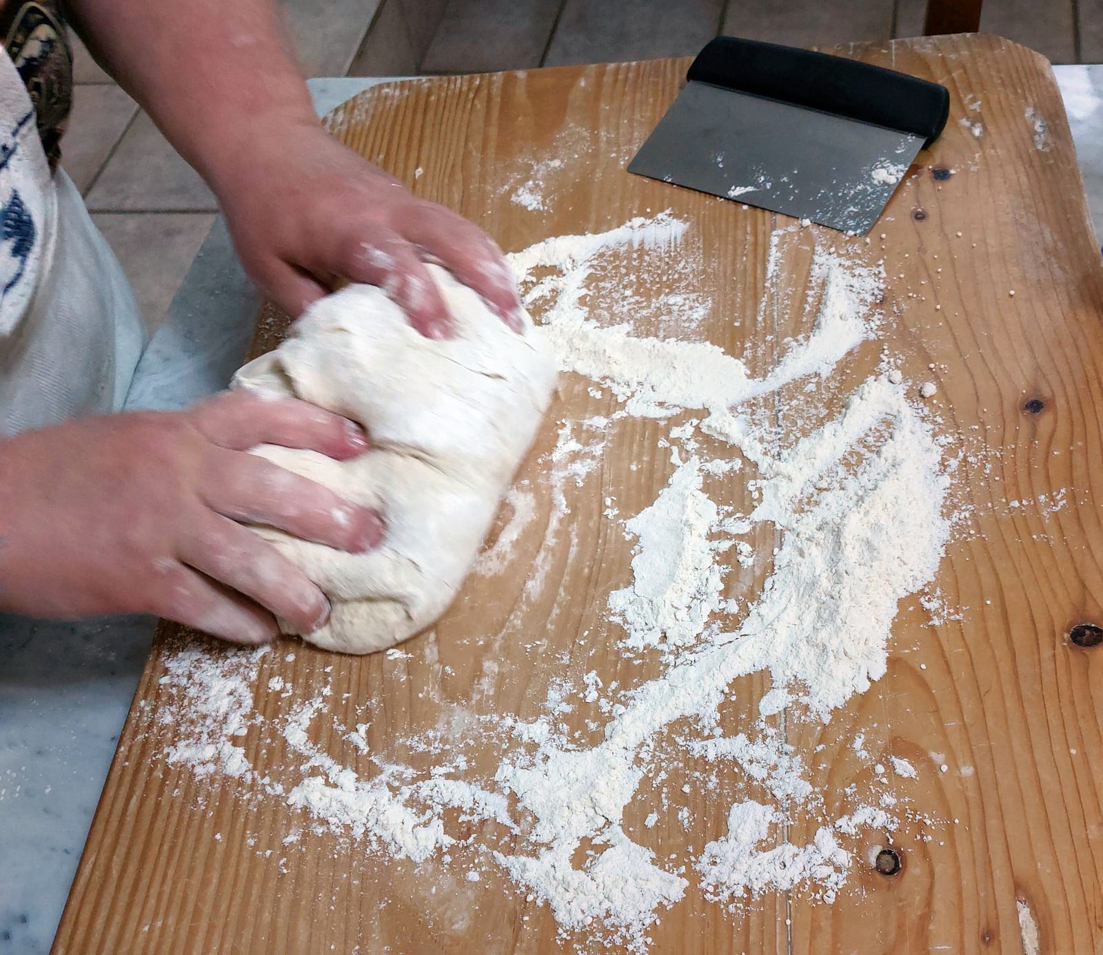 Hands kneading a round of bread dough on a floured wooden board.