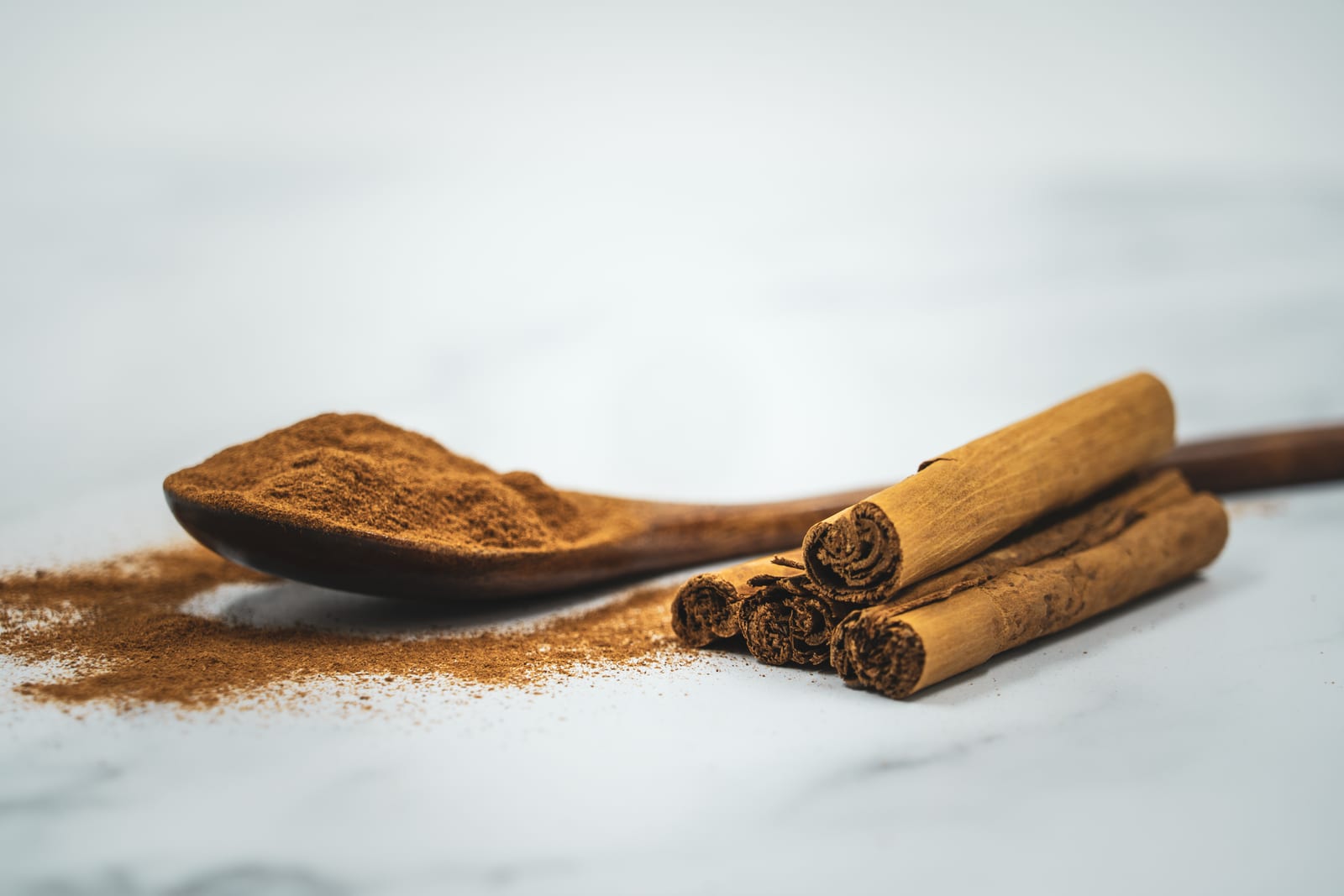 A spoonful of ground cinnamon beside curled cinnamon bark on a dark surface.
