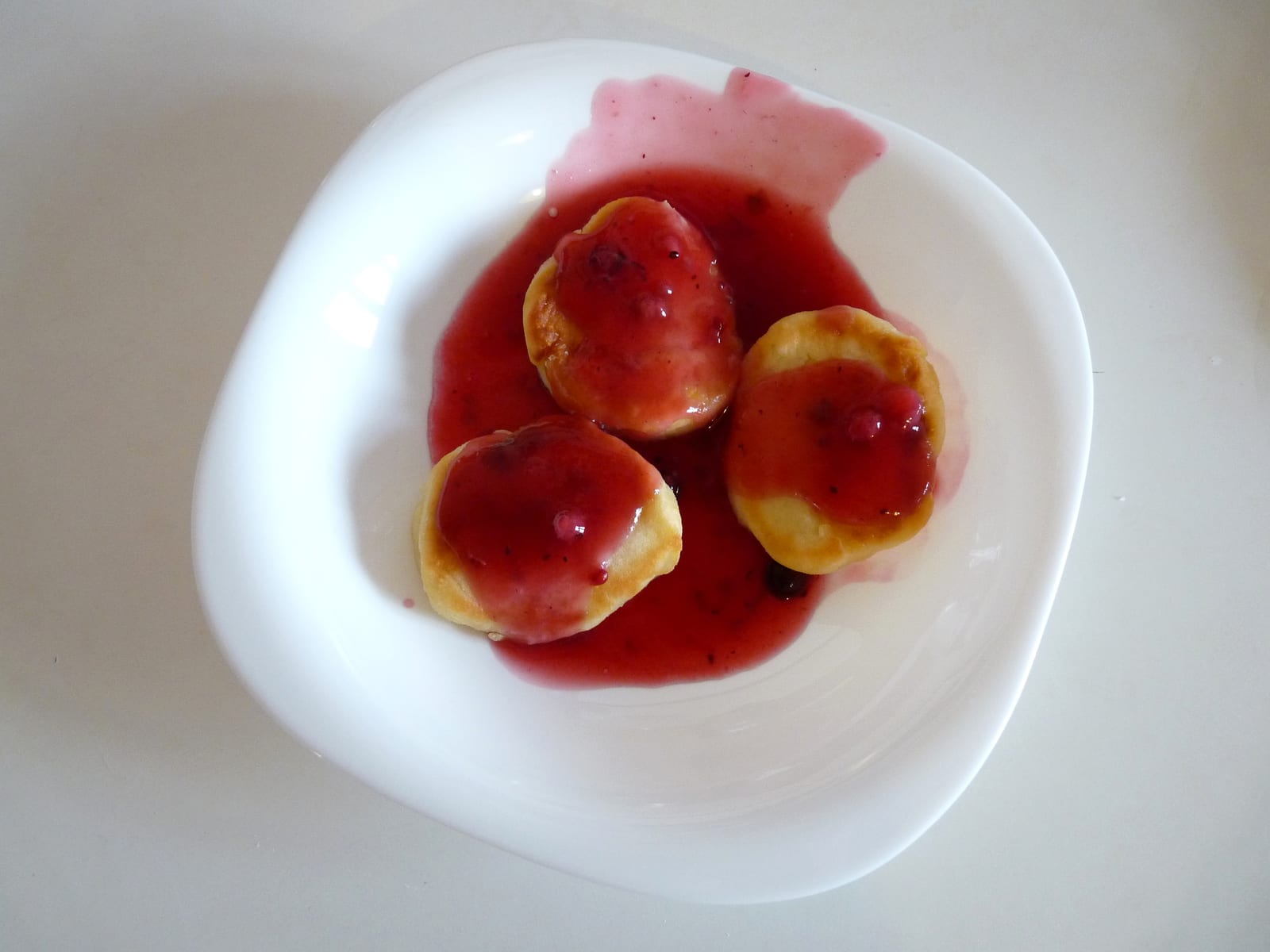 Three small golden syrniki covered with glossy red forest-berry kissel on a white plate.