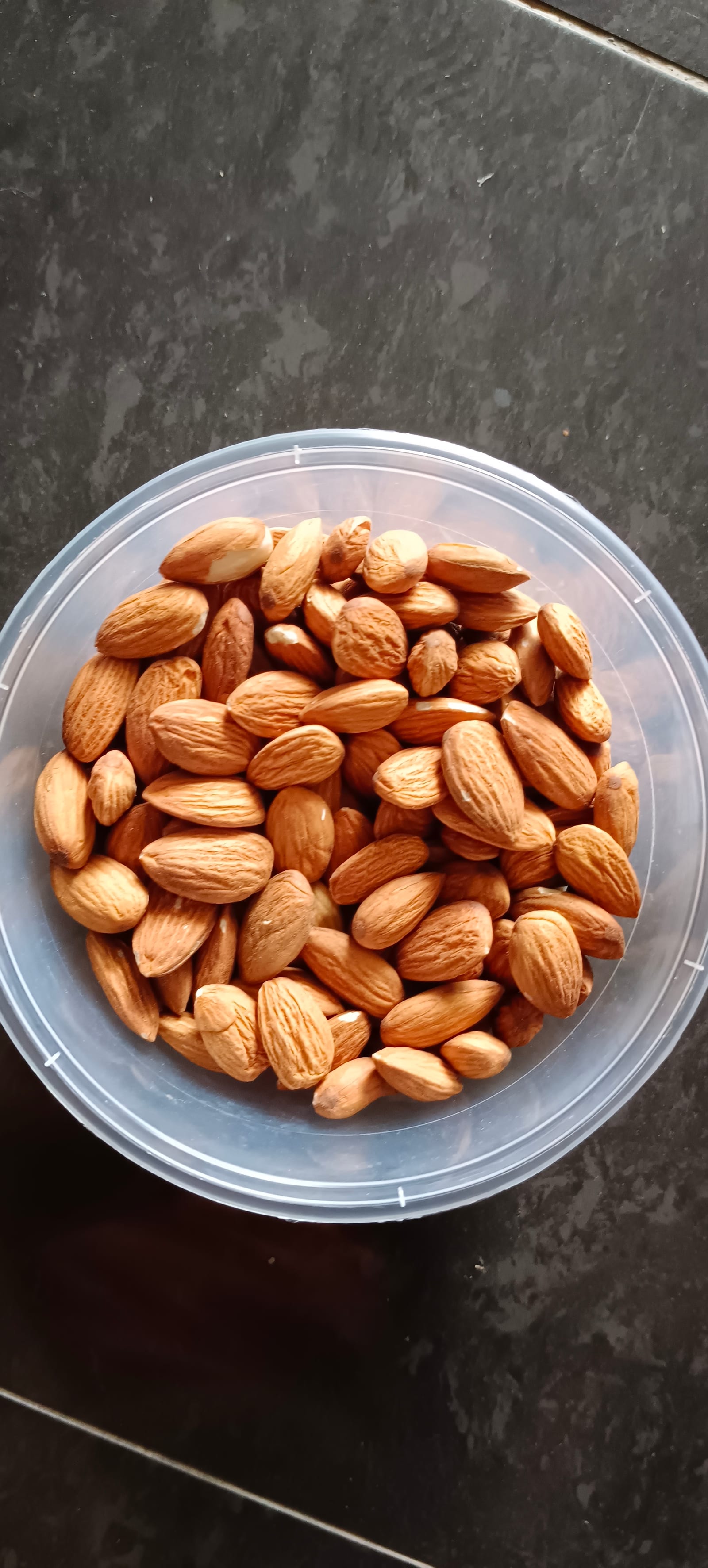 A clear bowl filled with whole brown-skinned almonds on a dark stone surface.